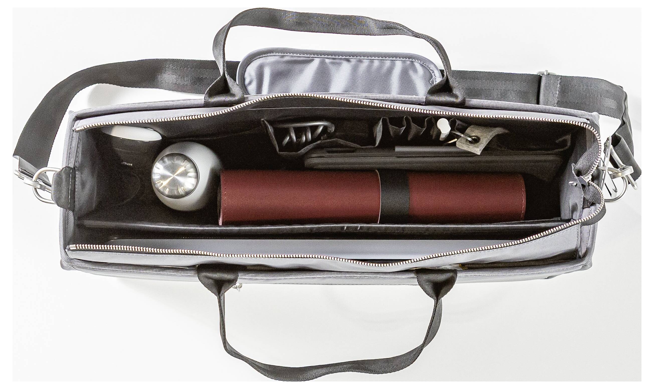 Eine Draufsicht eines geöffneten Aktenkoffers, der ein vollgepacktes Innenleben mit einem zylindrischen roten Etui, einem silbernen Thermobecher und verschiedenen Fächern zeigt.