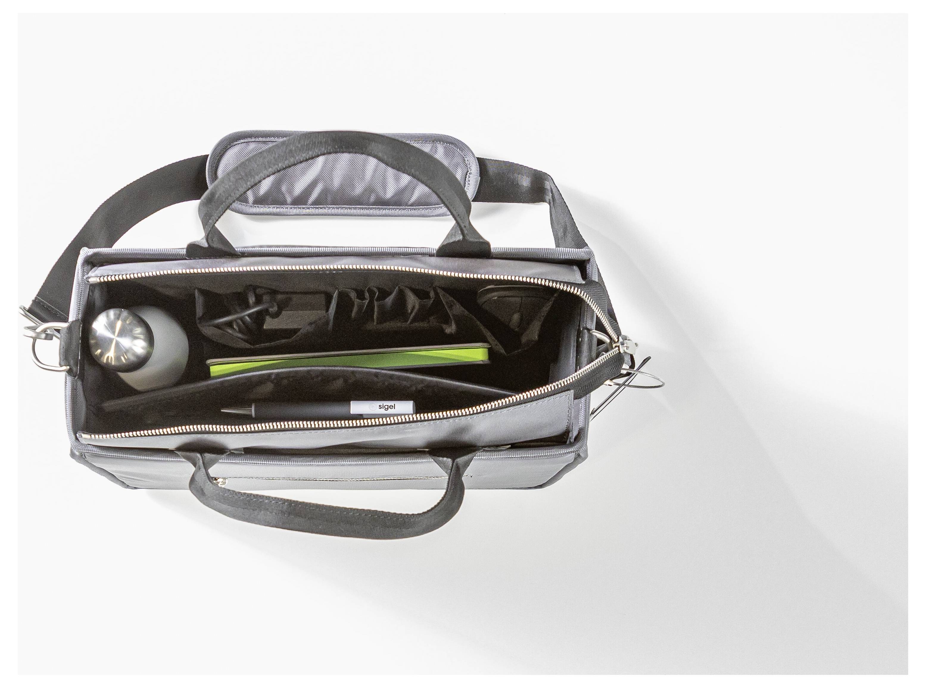 Schwarze Tasche von oben geöffnet, die eine Wasserflasche, einen schwarzen Beutel und ein langes, grünes Objekt enthält. Sie befindet sich auf einer weißen Oberfläche.