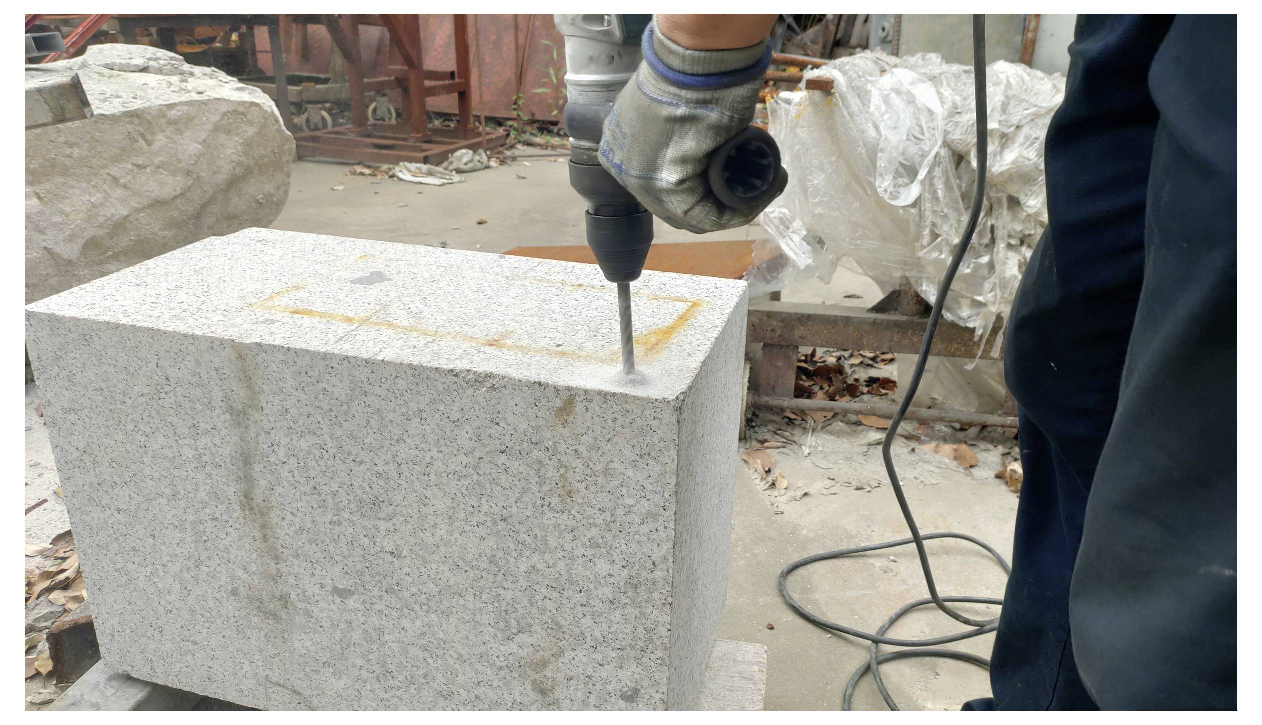 Eine Person benutzt eine Bohrmaschine an einem großen Steinblock in einer Freiluft-Werkstatt. Der Bereich ist gefüllt mit verschiedenen Werkzeugen und Materialien.