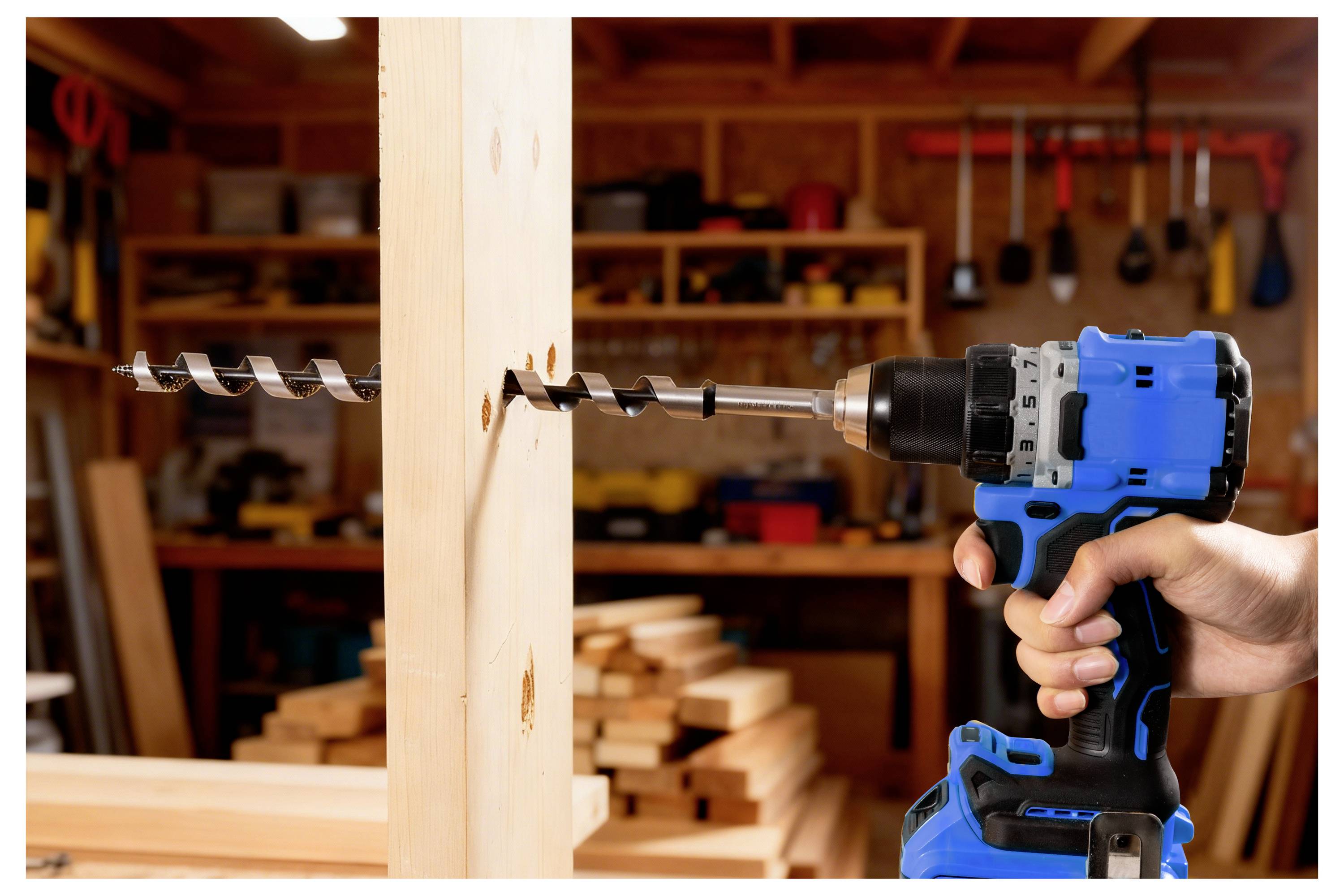 Eine Person verwendet eine blaue kabellose Bohrmaschine mit einem großen Bohrer, um Löcher in einen Holzbalken in einer Werkstatt mit Werkzeugen im Hintergrund zu bohren.