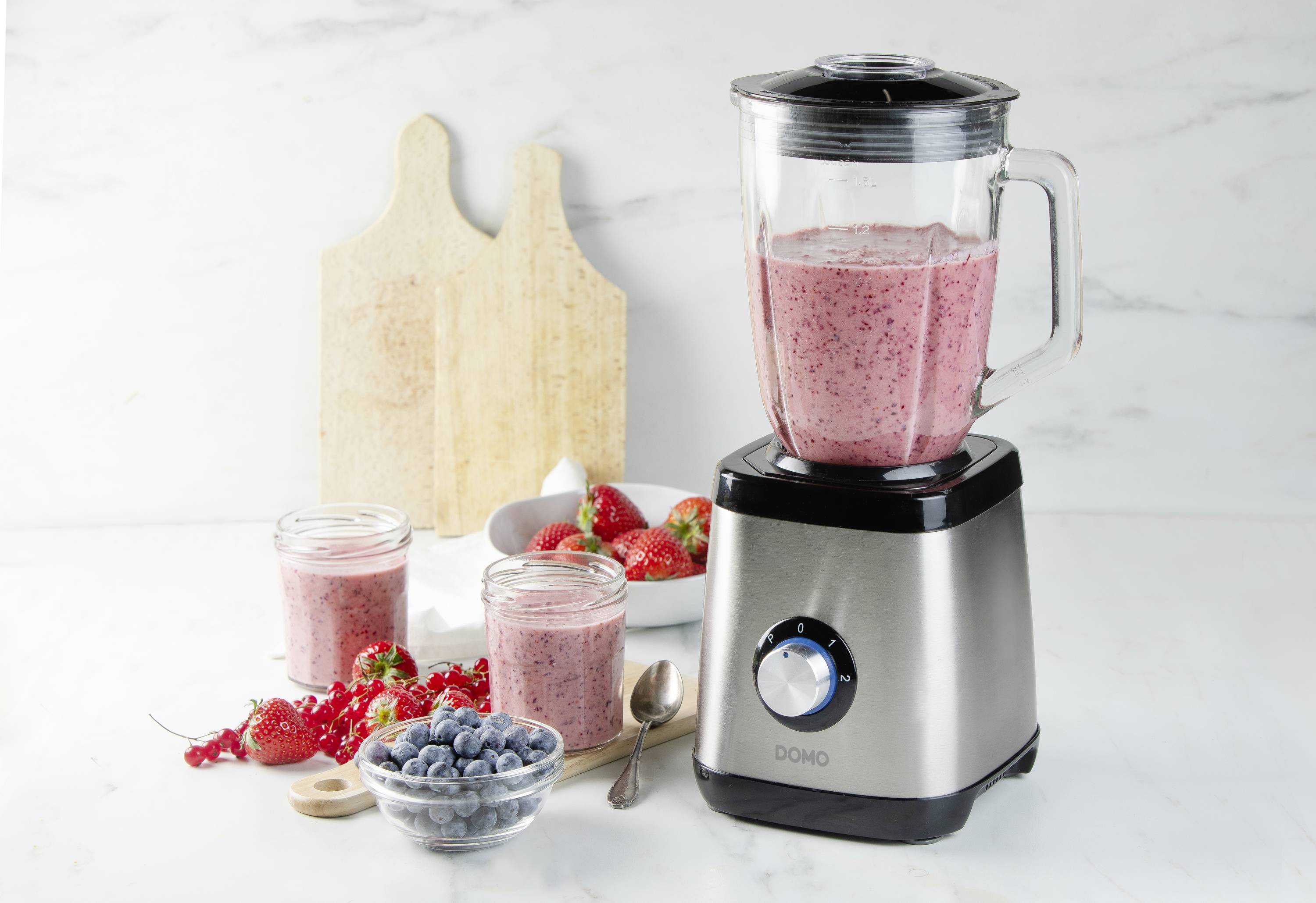 Ein Standmixer enthält roten Frucht-Smoothie. Daneben stehen gefüllte Gläser, frische Erdbeeren, Blaubeeren und ein Löffel.