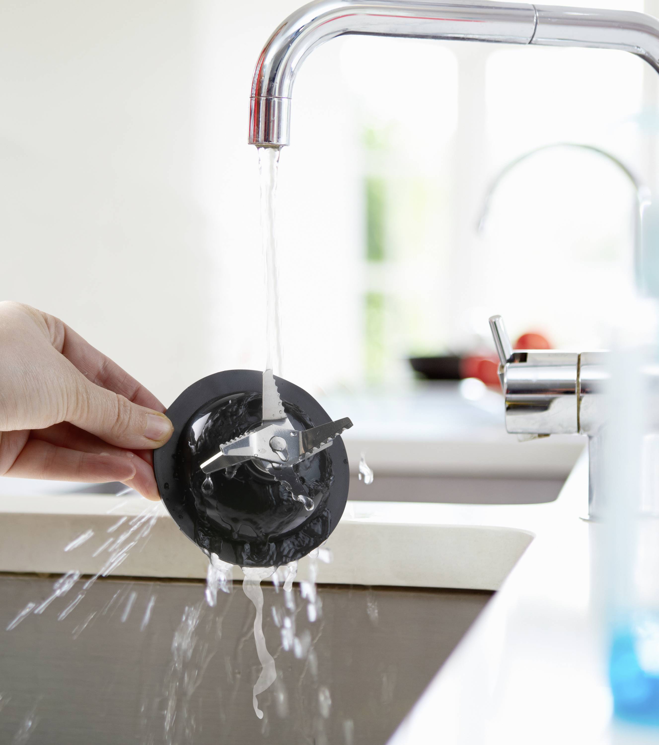 Eine Hand hält einen Abflussstopfen unter fließendem Wasser, um ihn zu reinigen. Im Hintergrund ist ein unscharfes Fenster zu sehen.