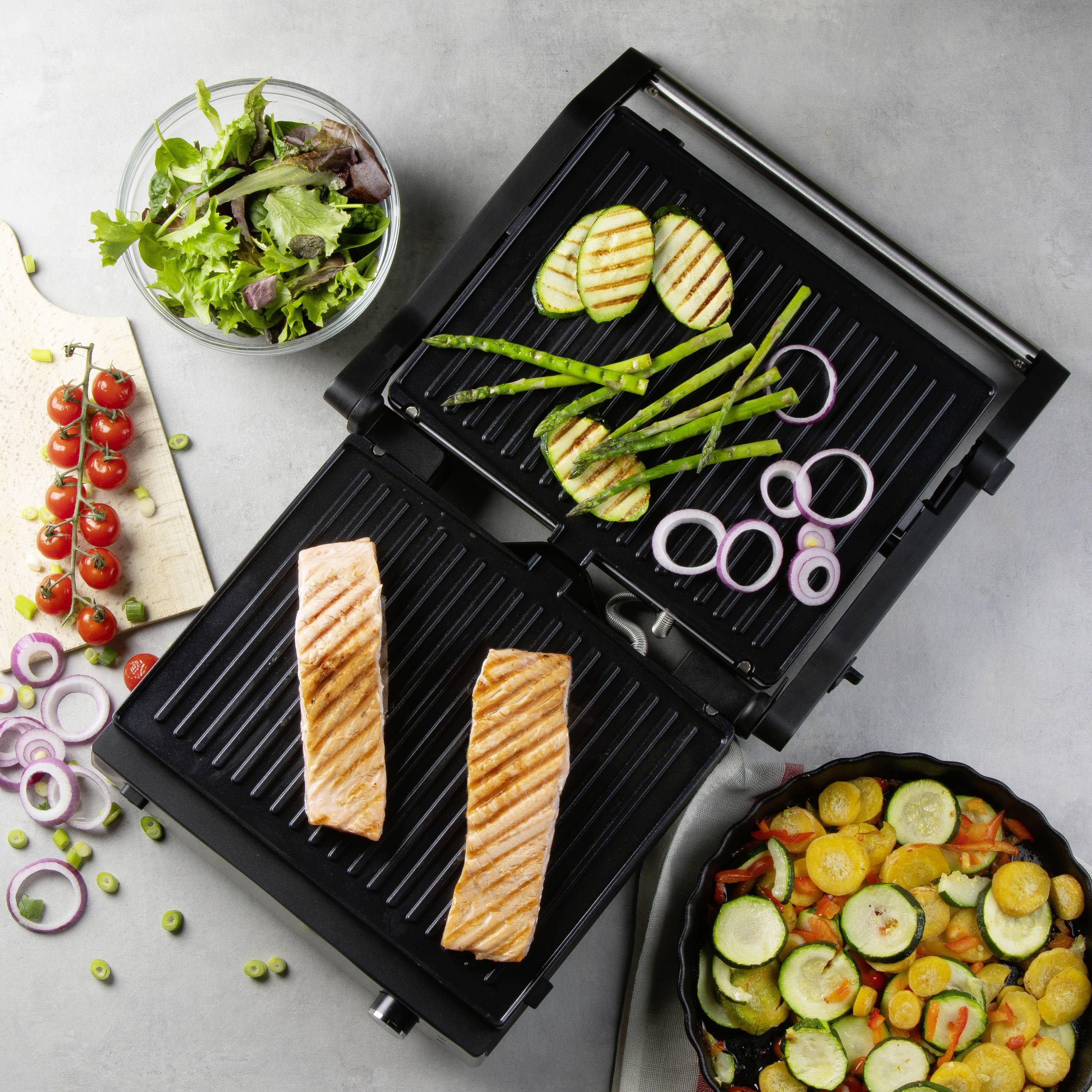 Ein Elektrogrill mit zwei Lachsfilets brät, umgeben von frischem Gemüse wie Zucchini, Tomaten und Salat in Schalen und auf Brettern.