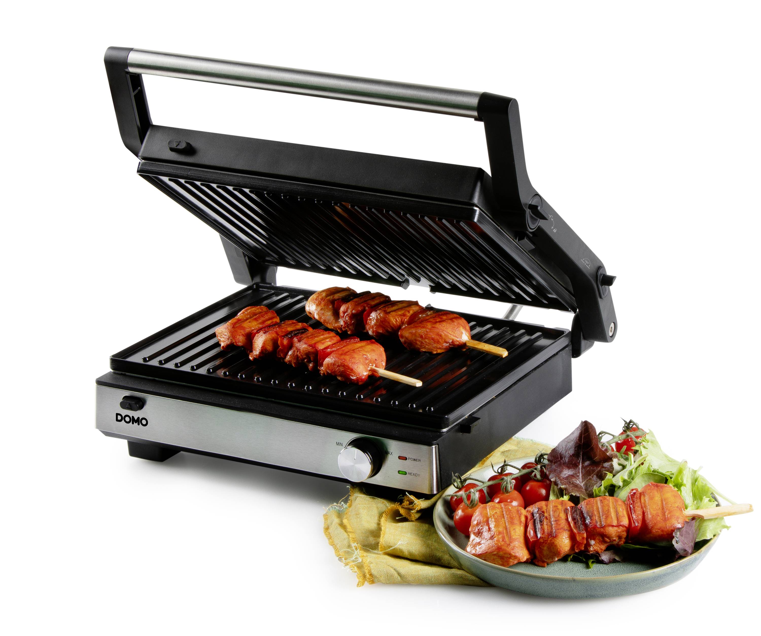 Ein Elektrogrill mit geöffnetem Deckel grillt zwei Fleischspieße. Daneben liegt ein Teller mit frischen Salatblättern und Tomaten.