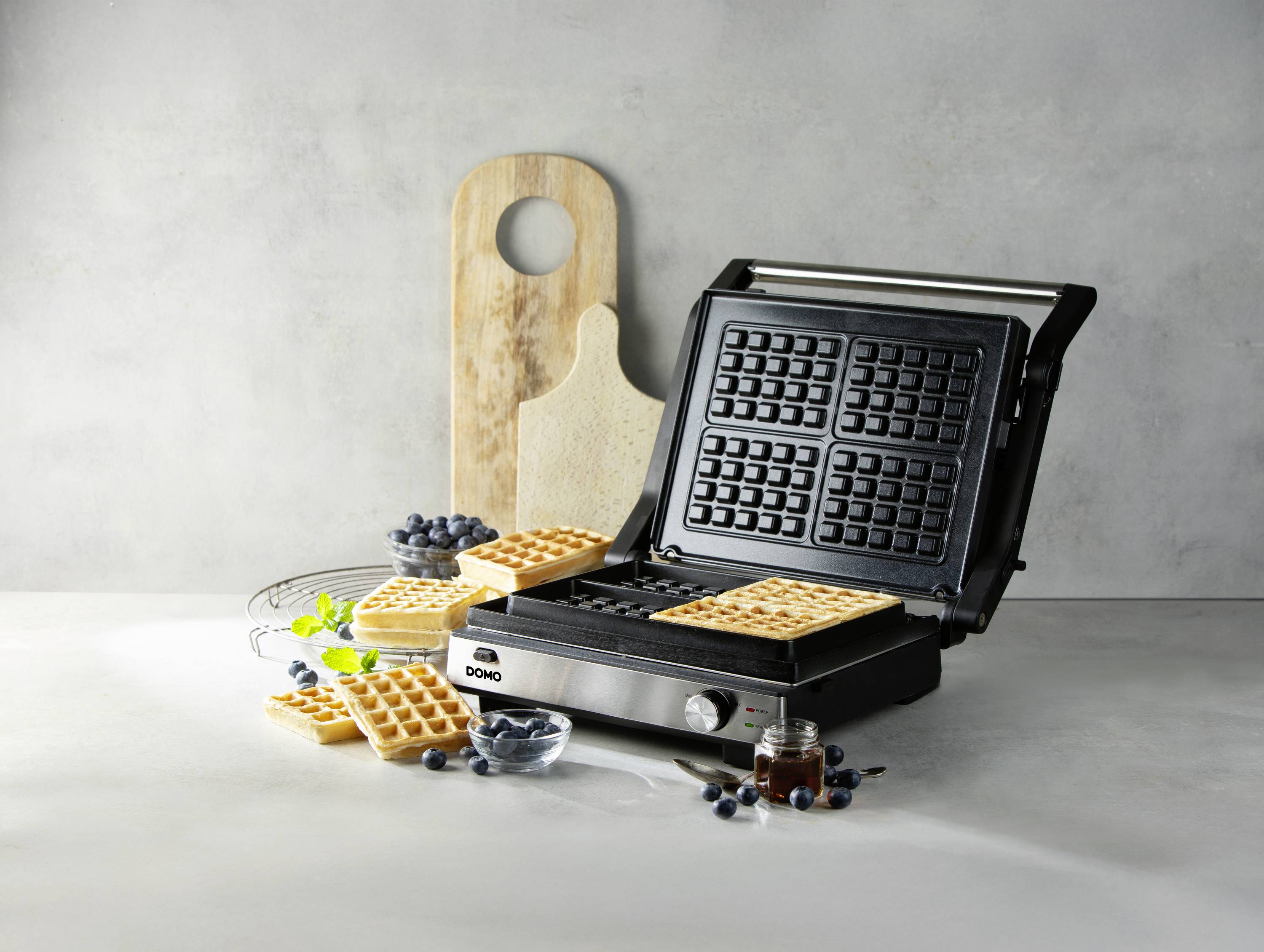 Waffeleisen in geöffneter Position mit frisch gebackenen Waffeln, umgeben von Heidelbeeren auf einem hellen Hintergrund.