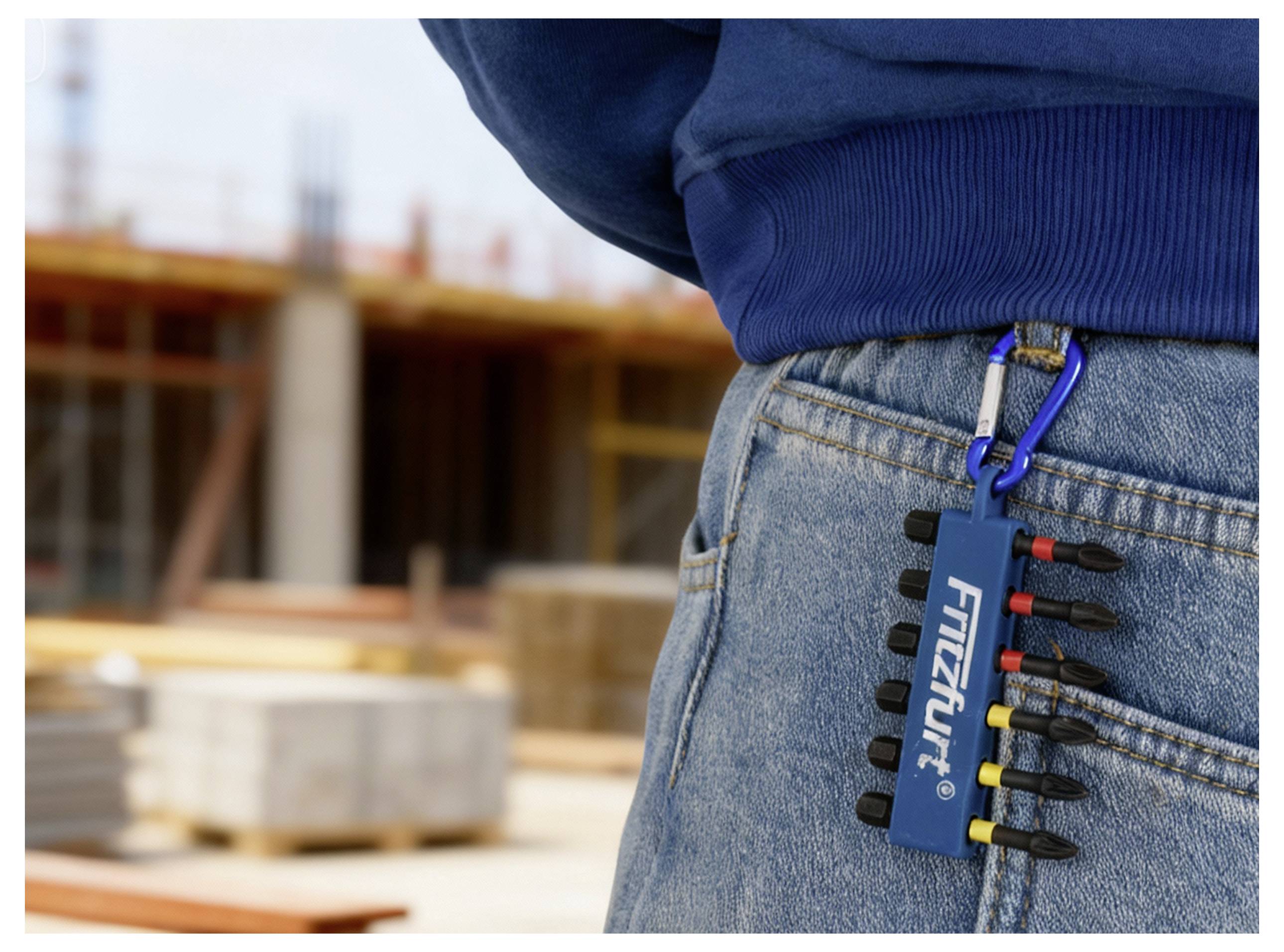 Eine Person trägt Jeans mit einem blauen Multitool, das an einem Karabiner befestigt ist, und steht auf einer Baustelle mit unscharf im Hintergrund liegenden Materialien.