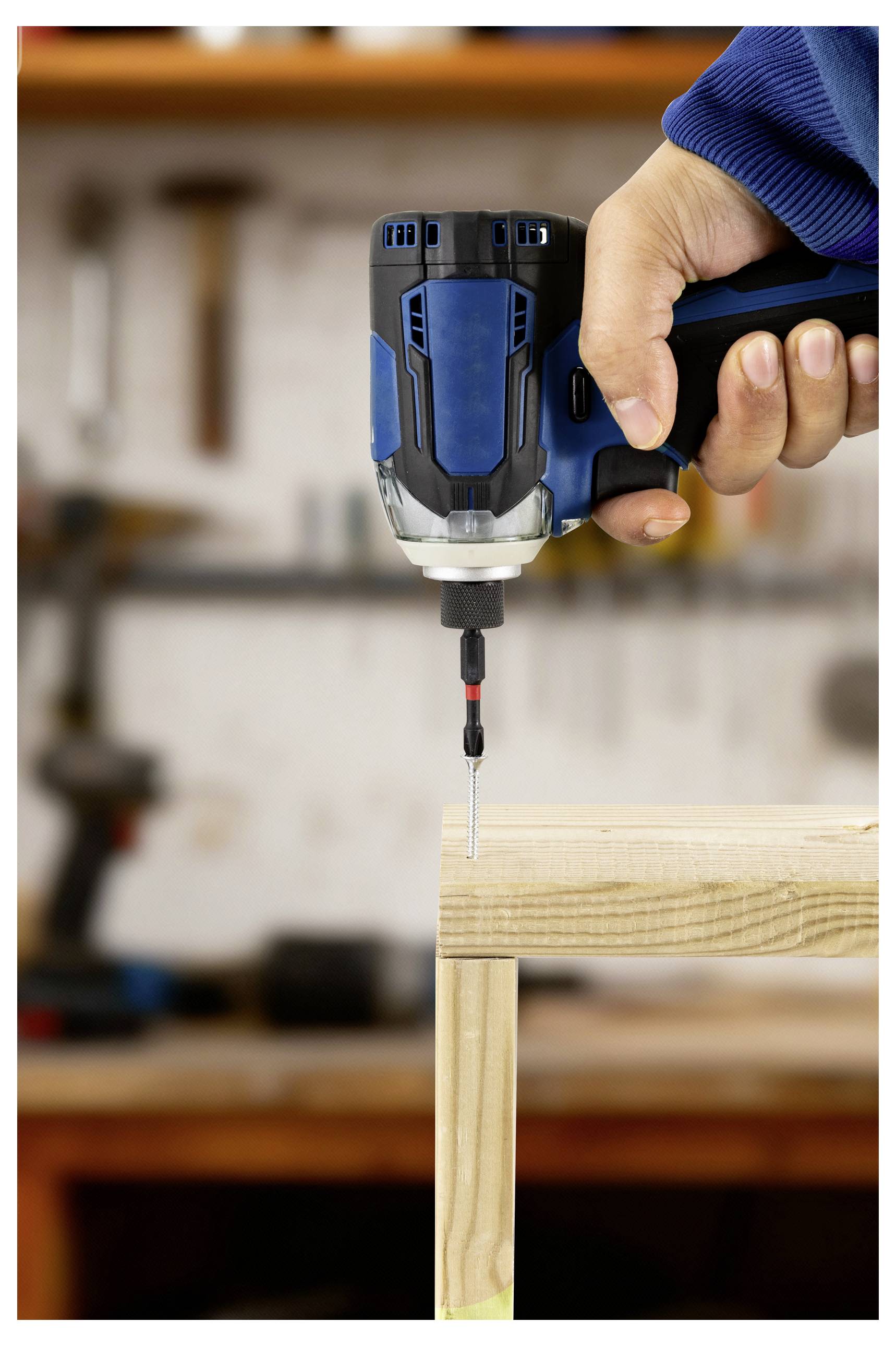 Eine Person verwendet einen blauen Akkuschrauber, um eine Schraube in einen Holzrahmen in einer Werkstattumgebung einzudrehen.