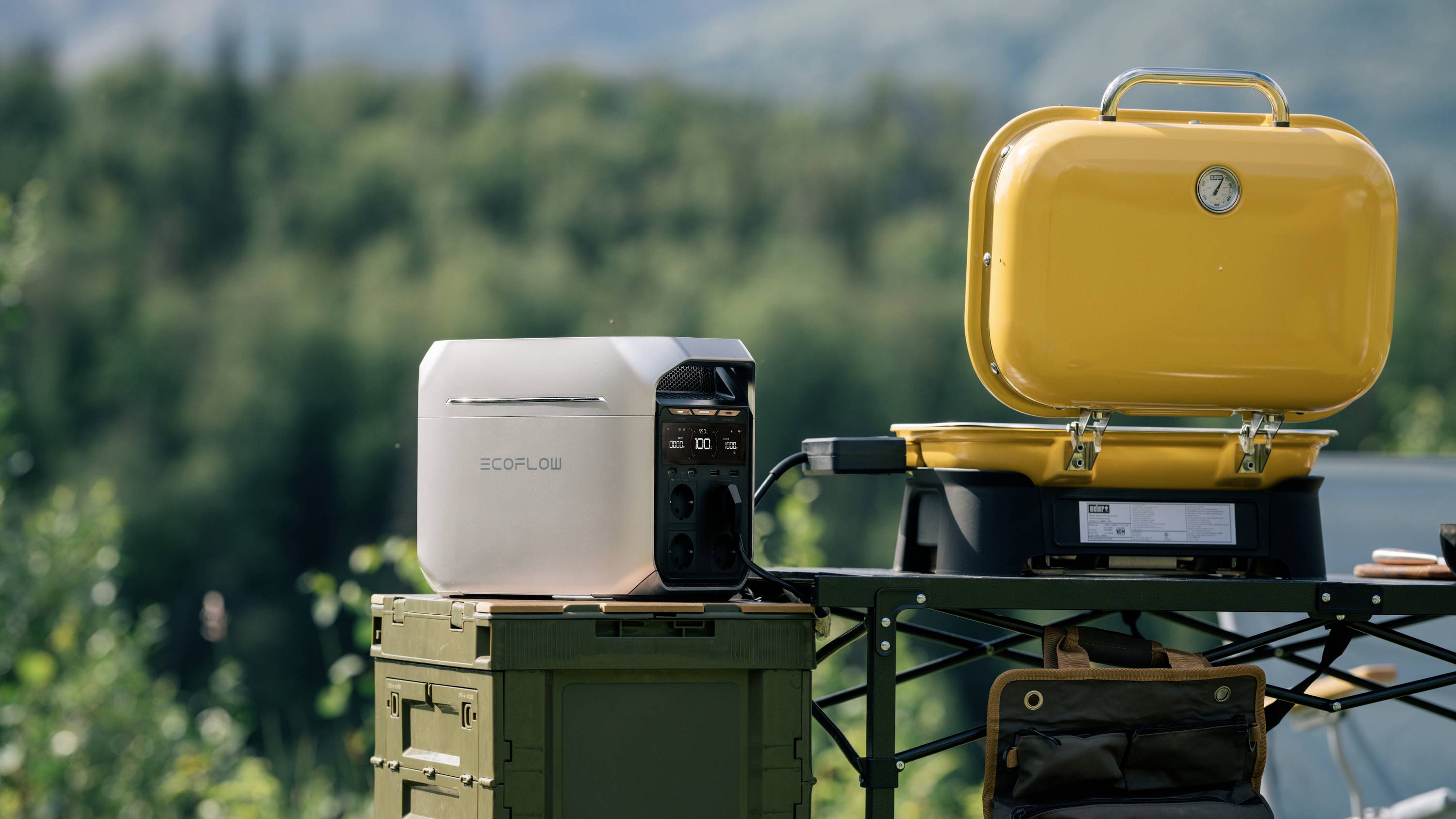 Ein tragbarer Stromgenerator steht neben einem gelben Grill auf einem Tisch im Freien vor einer verschwommenen Waldkulisse.