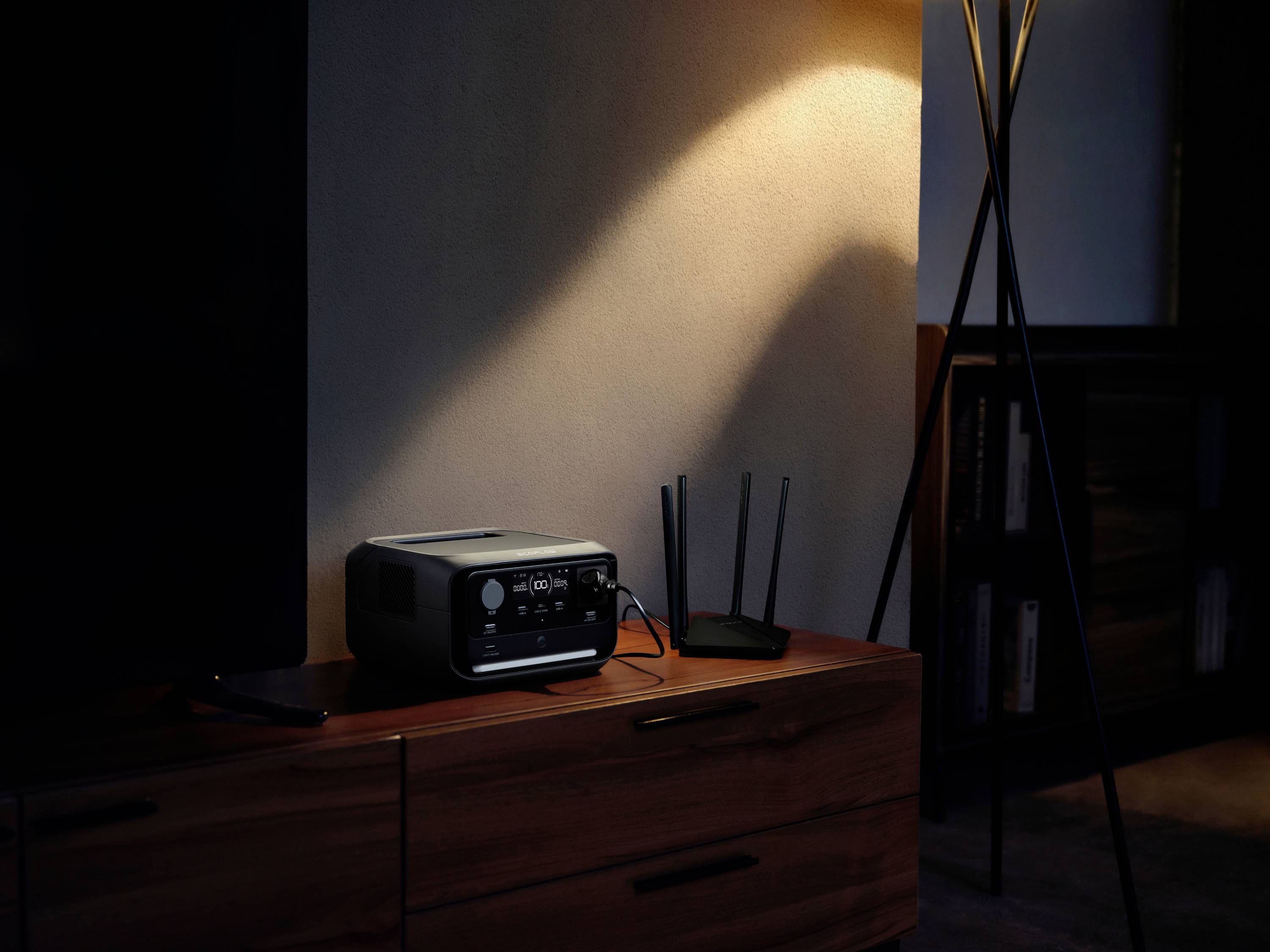 Ein Router und ein technisches Gerät stehen auf einem Holzschrank, beleuchtet von einer Stehlampe. Links ist ein Teil eines Bildschirms sichtbar.