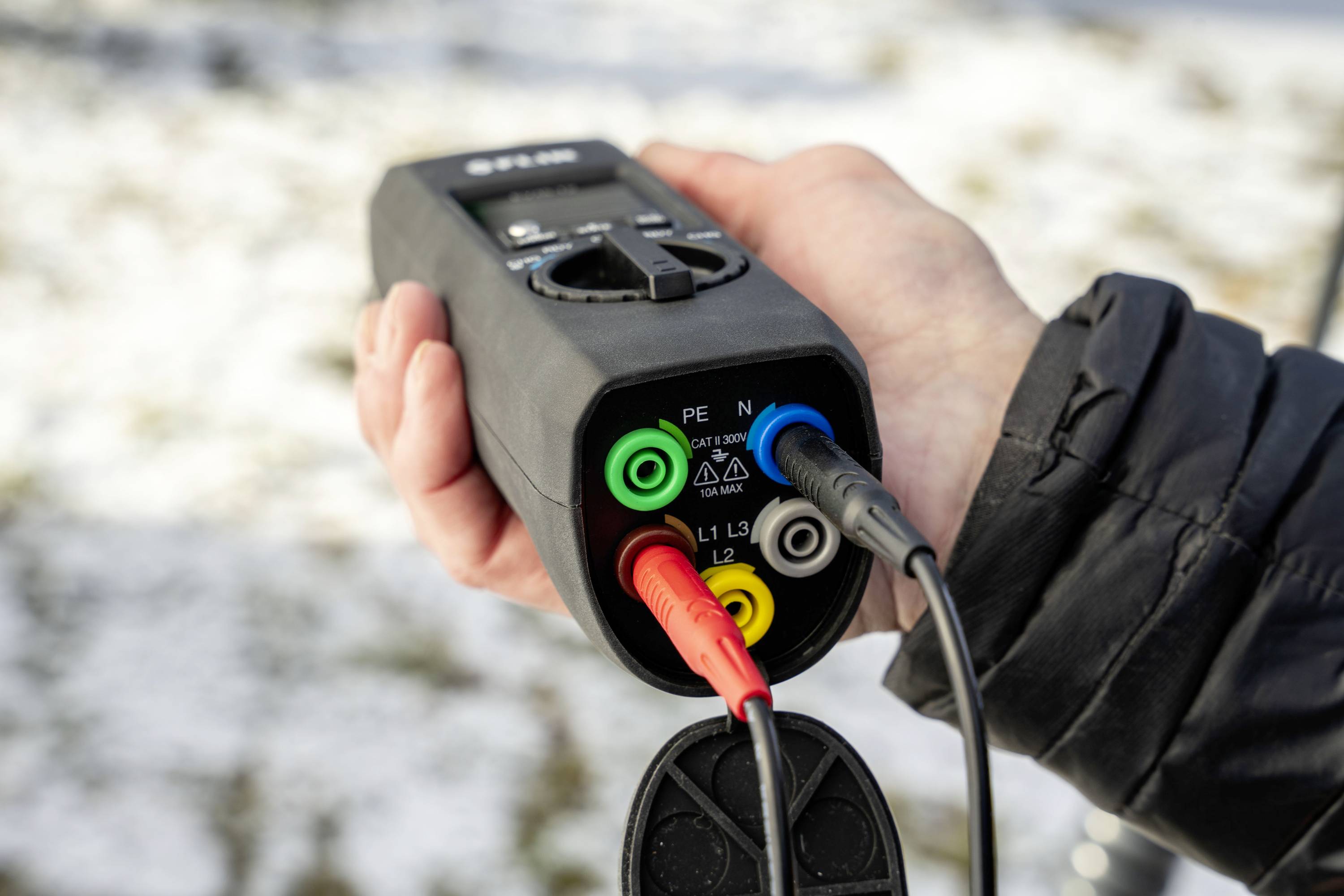 Eine Person hält ein elektrisches Prüfgerät mit bunten Kabeln, verwendet für Sicherheits- und Funktionstests von elektrischen Systemen. Im Hintergrund Schnee auf Gras.