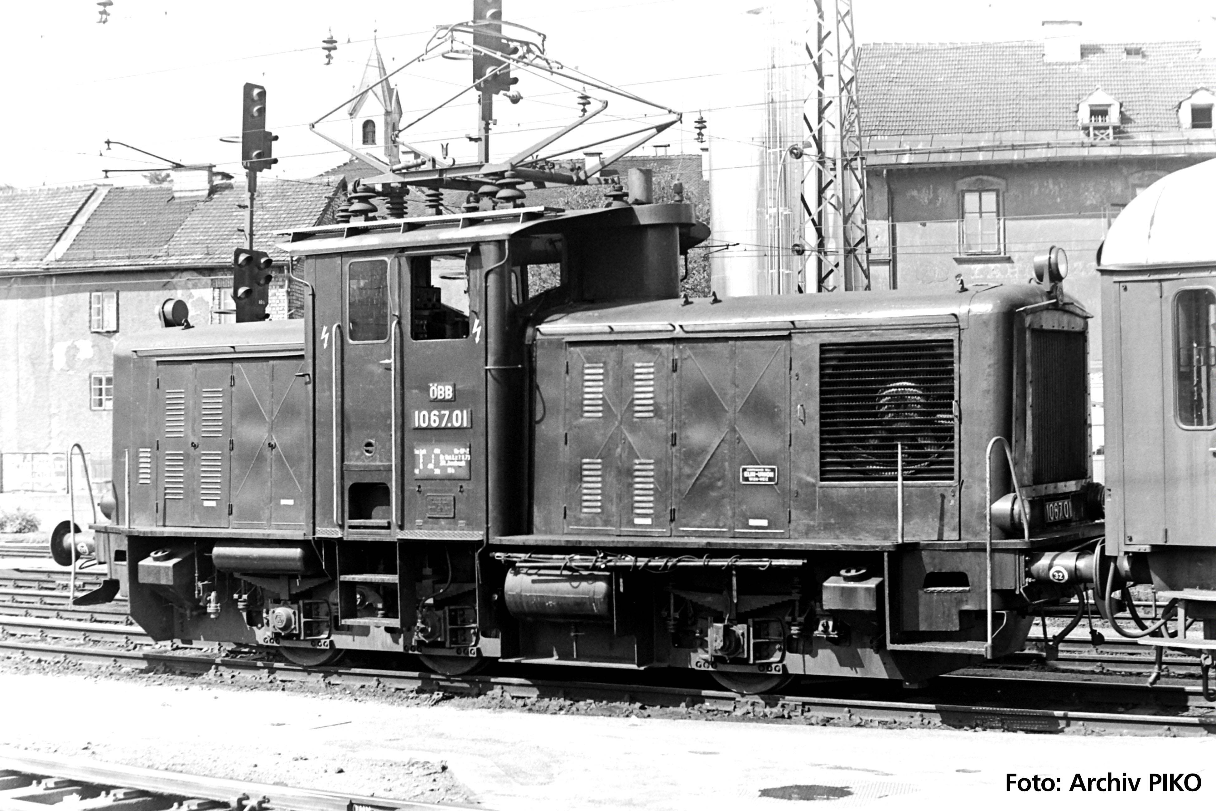 Schwarz-weiß Foto einer historischen Diesellokomotive auf Gleisen im Bahnhofsbereich. Im Hintergrund sind Gebäude zu sehen.