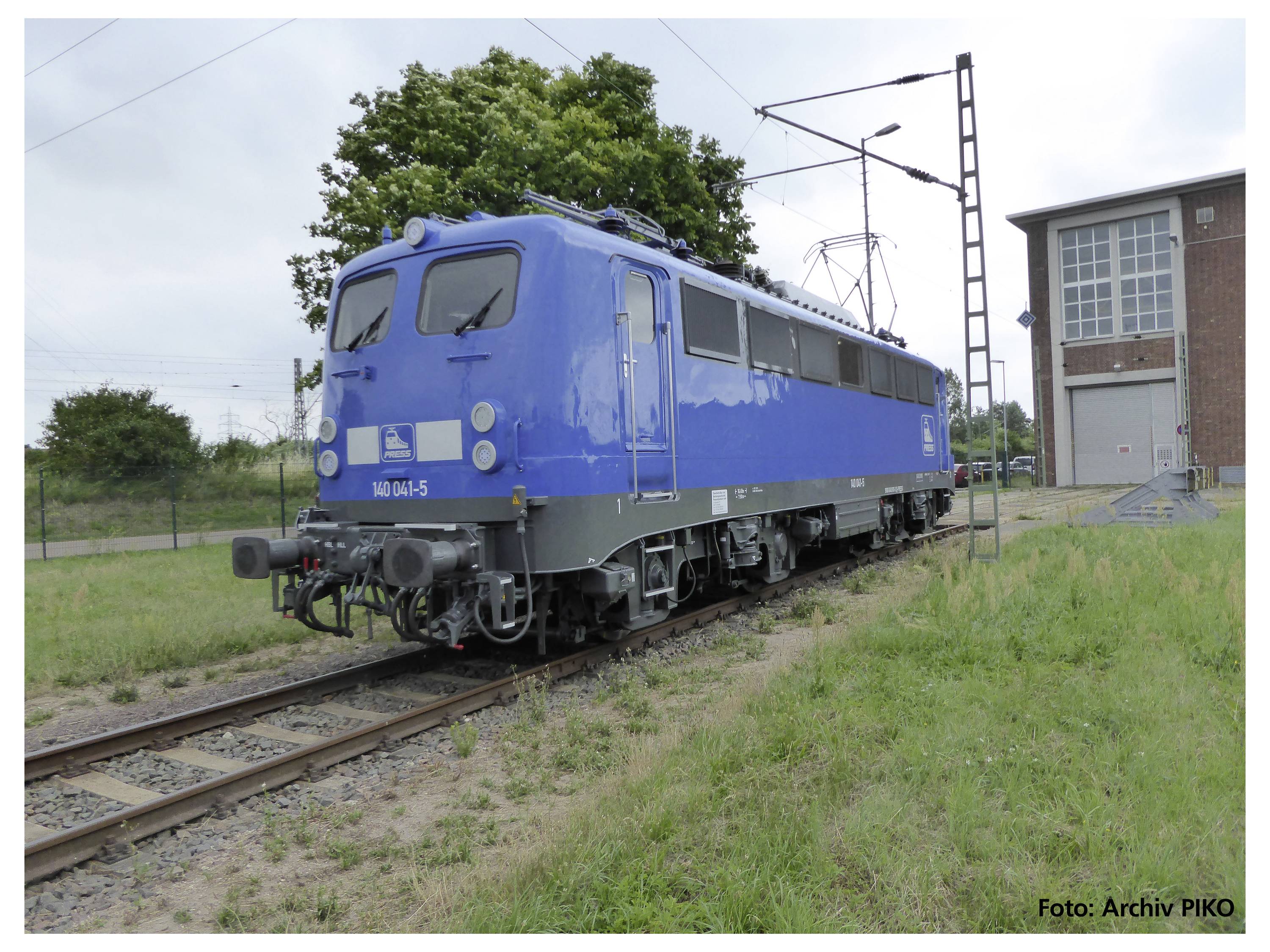 Eine blaue Elektrolokomotive steht auf einem Gleis vor einem Gebäude. Im Hintergrund sind Bäume und Stromleitungen zu sehen.