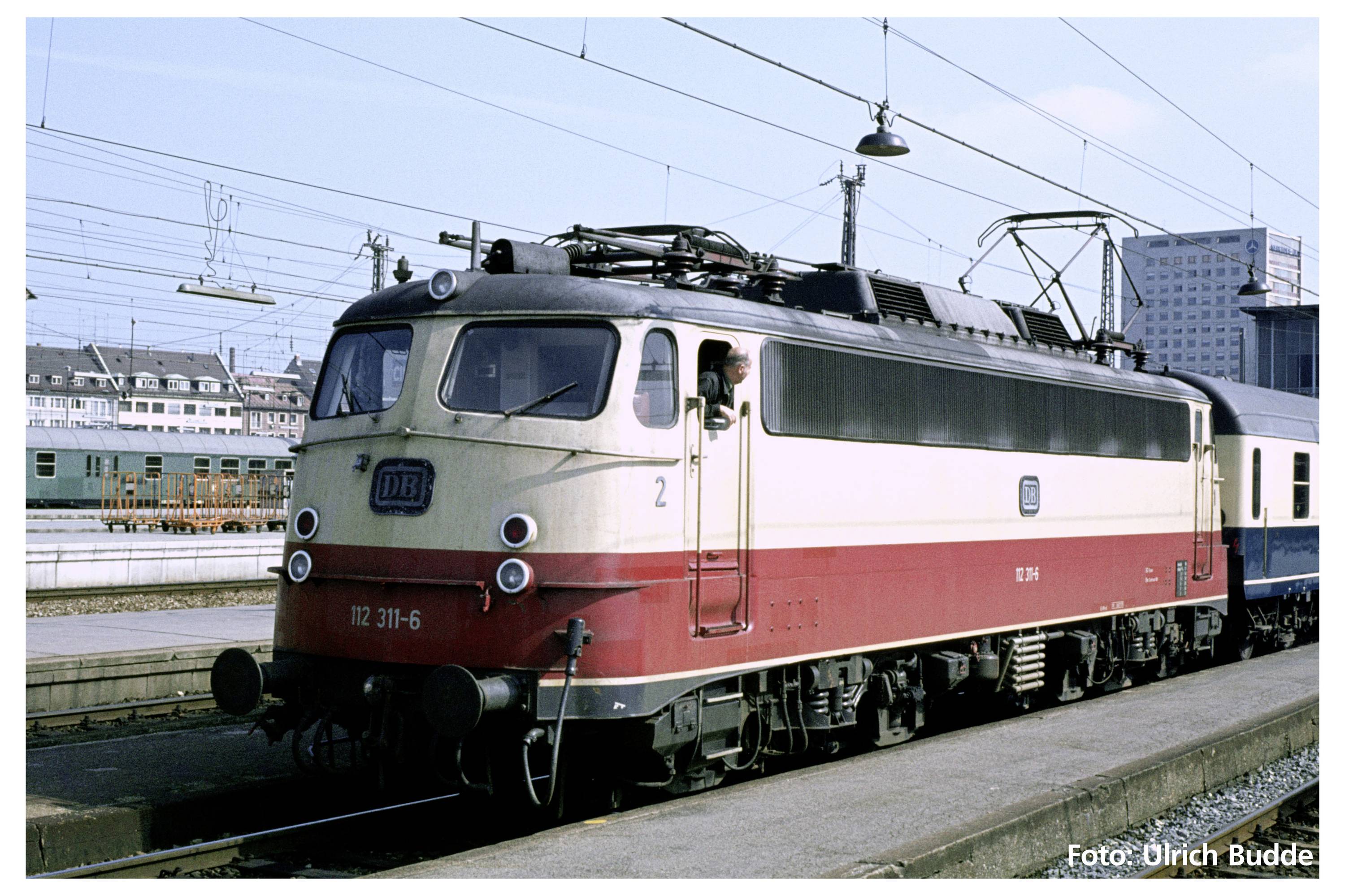 Eine rote und cremefarbene Lokomotive steht auf einem Bahnhofsgleis. Gebäudestrukturen und elektrische Leitungen im Hintergrund.