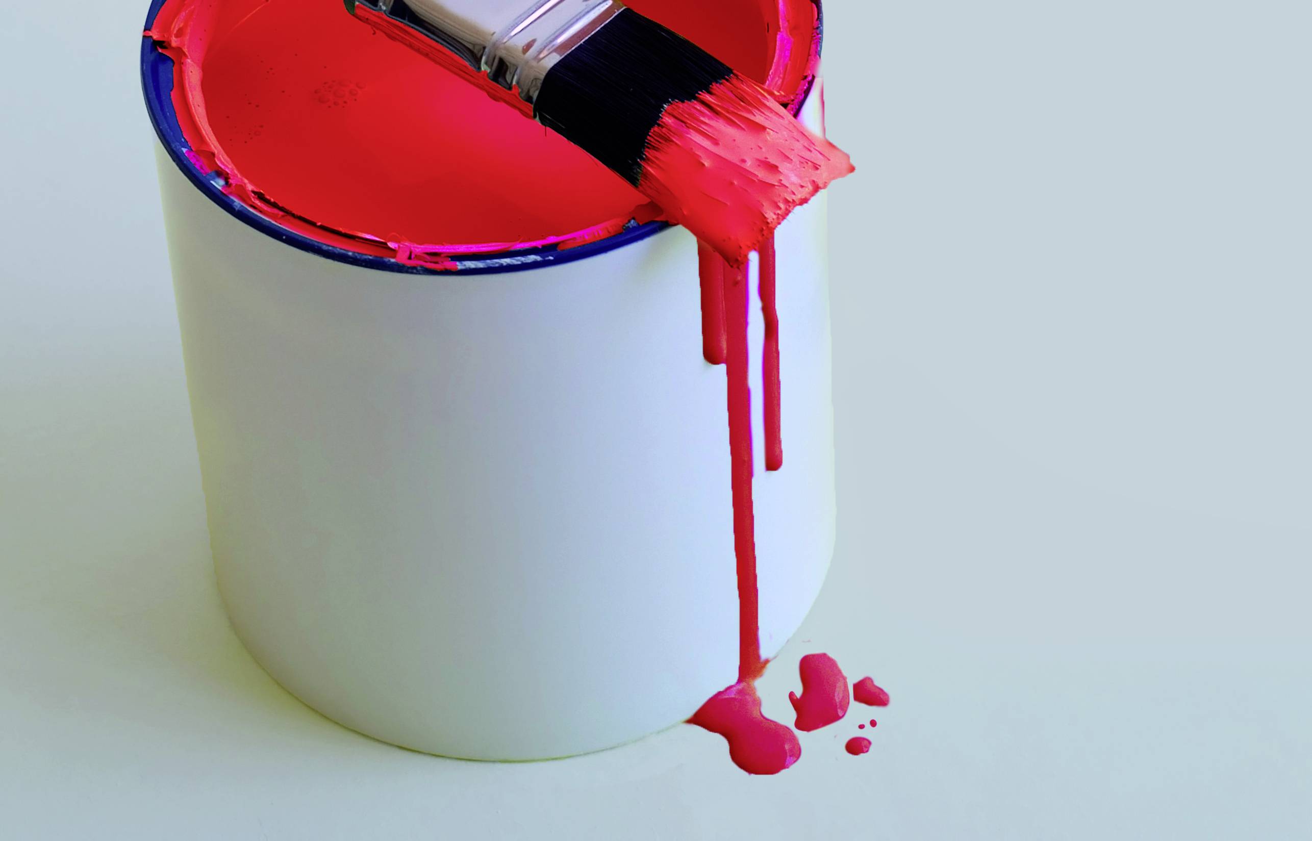 Ein offener Farbkübel mit roter Farbe, ein Pinsel liegt diagonal darauf und rote Farbe tropft den Kübel seitlich herunter.