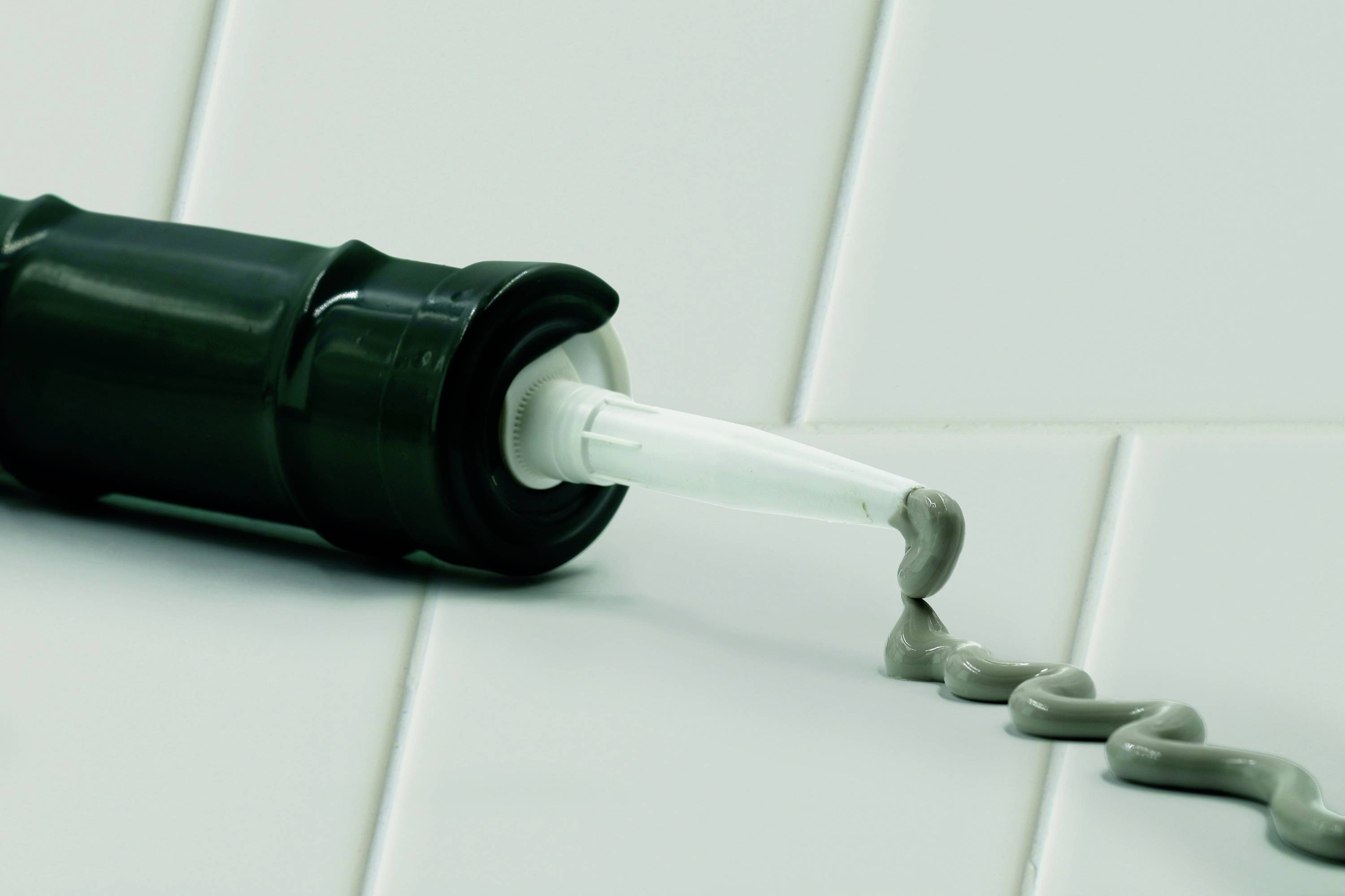 Eine schwarze Kartuschenpistole liegt auf weißen Fliesen und gibt eine graue Abdichtungsmasse ab, die in einer S-förmigen Linie verläuft.