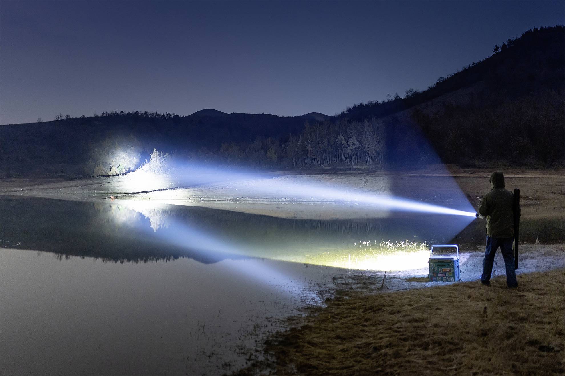 Eine Person steht am Ufer eines Sees und leuchtet mit einer starken Taschenlampe auf das gegenüberliegende Ufer in der Nacht.