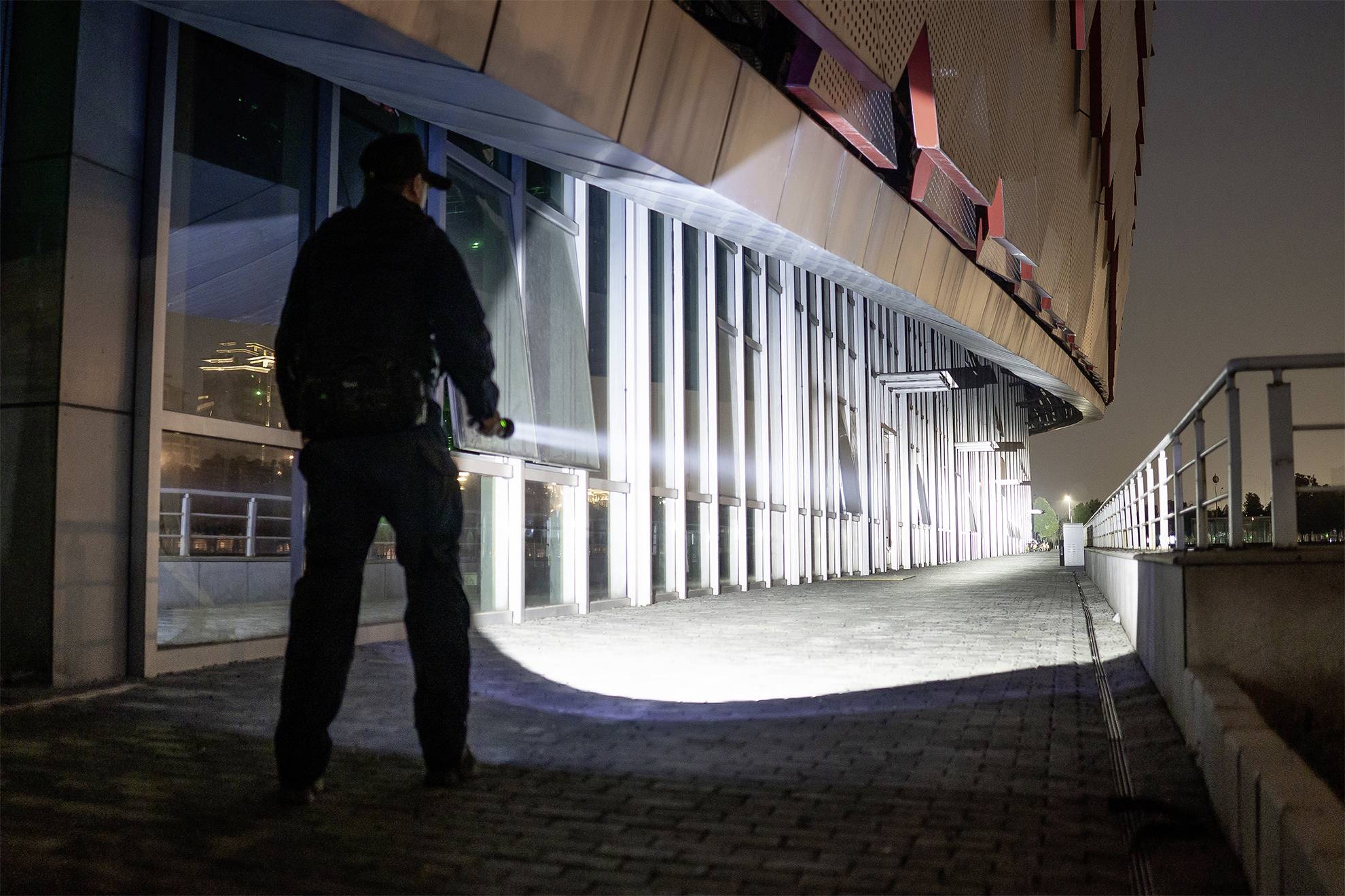 Eine Person in Uniform leuchtet nachts mit einer Taschenlampe auf den Boden vor einem modernen Gebäude.
