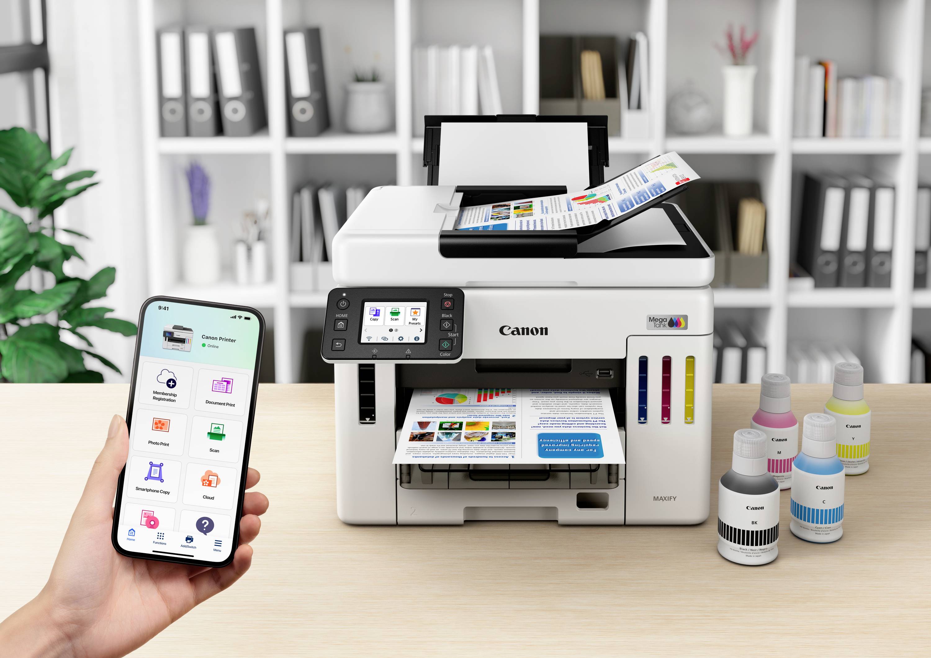Eine Person bedient einen Drucker per Smartphone. Der Drucker auf einem Tisch druckt farbige Dokumente. Rechts daneben stehen Tintenflaschen.