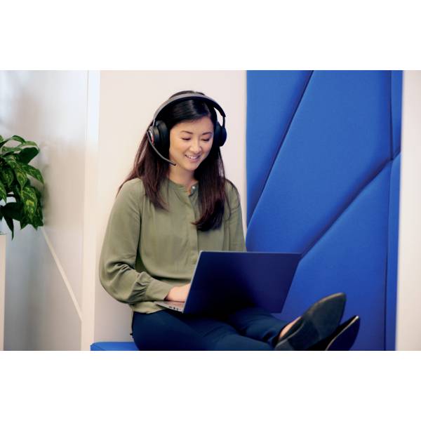 Frau mit Kopfhörern lächelt und arbeitet an einem Laptop, sitzt auf blauer Bank mit Pflanze im Hintergrund.