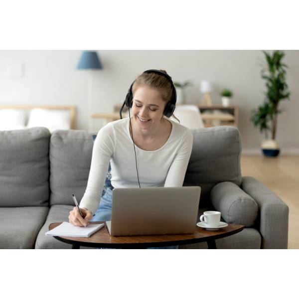 Frau sitzt auf einem Sofa, trägt Kopfhörer und schreibt Notizen, während sie auf einen Laptop schaut. Im Hintergrund ist eine gemütliche Wohnzimmereinrichtung zu sehen.