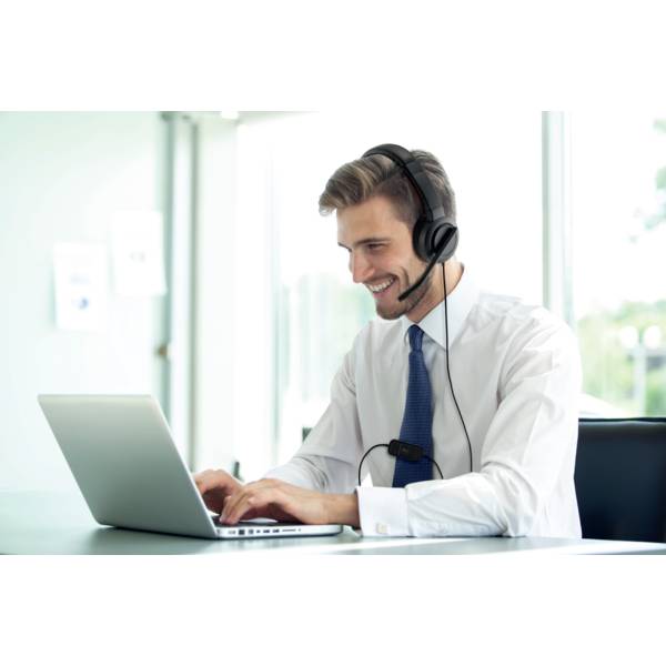 Ein lächelnder Mann trägt ein Headset und arbeitet an einem Laptop in einem modernen Büro. Im Hintergrund sind Fenster zu sehen.