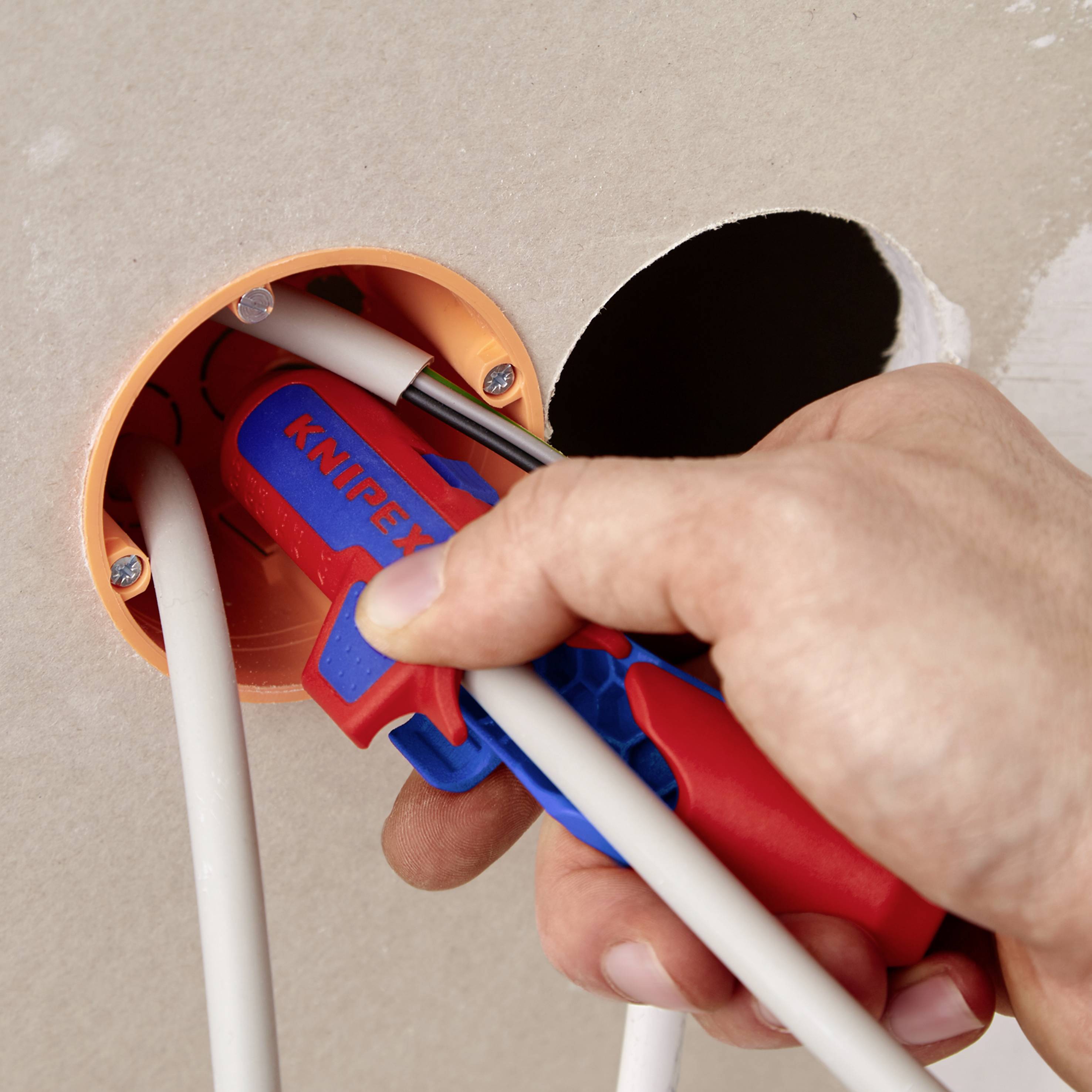 Eine Hand hält ein Knipex-Werkzeug und schneidet ein Kabel innerhalb einer Wandöffnung.