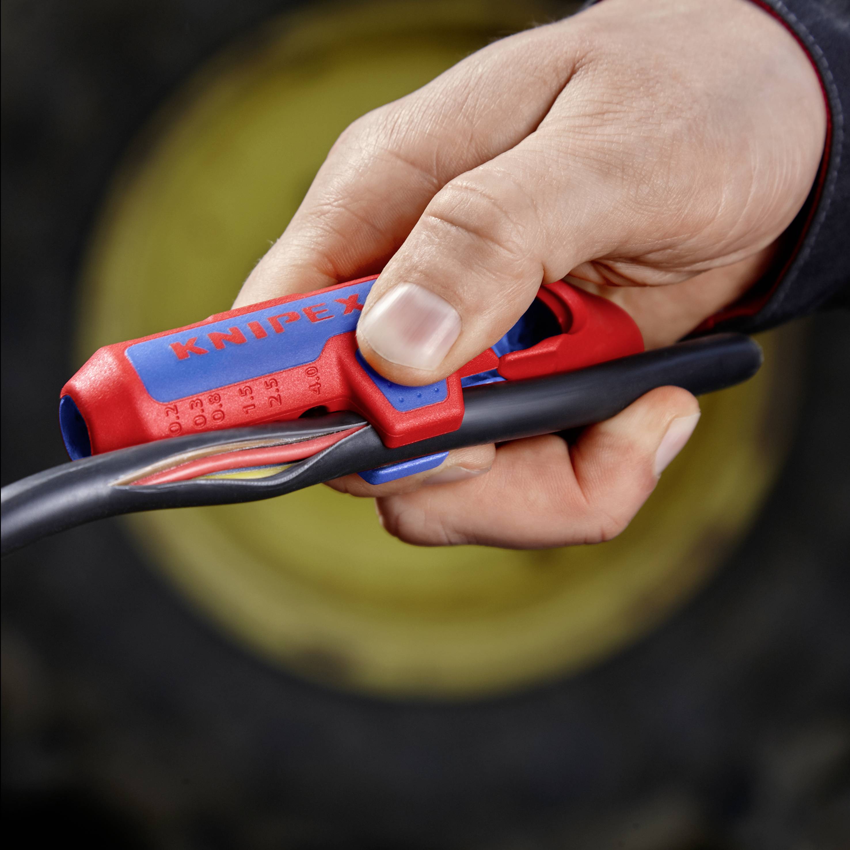 Eine Hand hält ein rotes Schneidewerkzeug, das auf einem schwarzen Kabel positioniert ist, vor einem unscharfen, runden gelben Hintergrund.