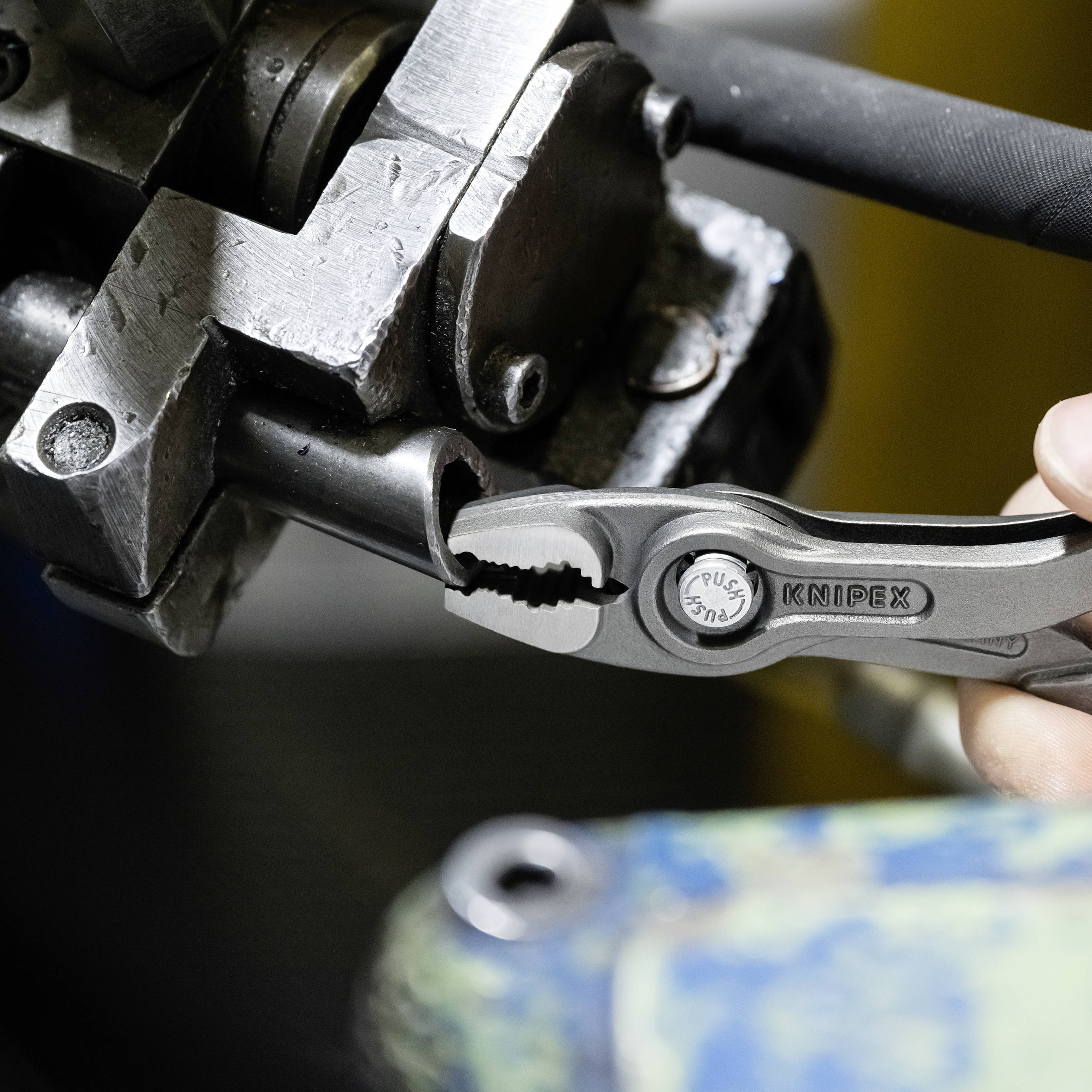 Eine Hand hält eine KNIPEX-Zange und greift ein Metallteil in einer Werkstattumgebung.