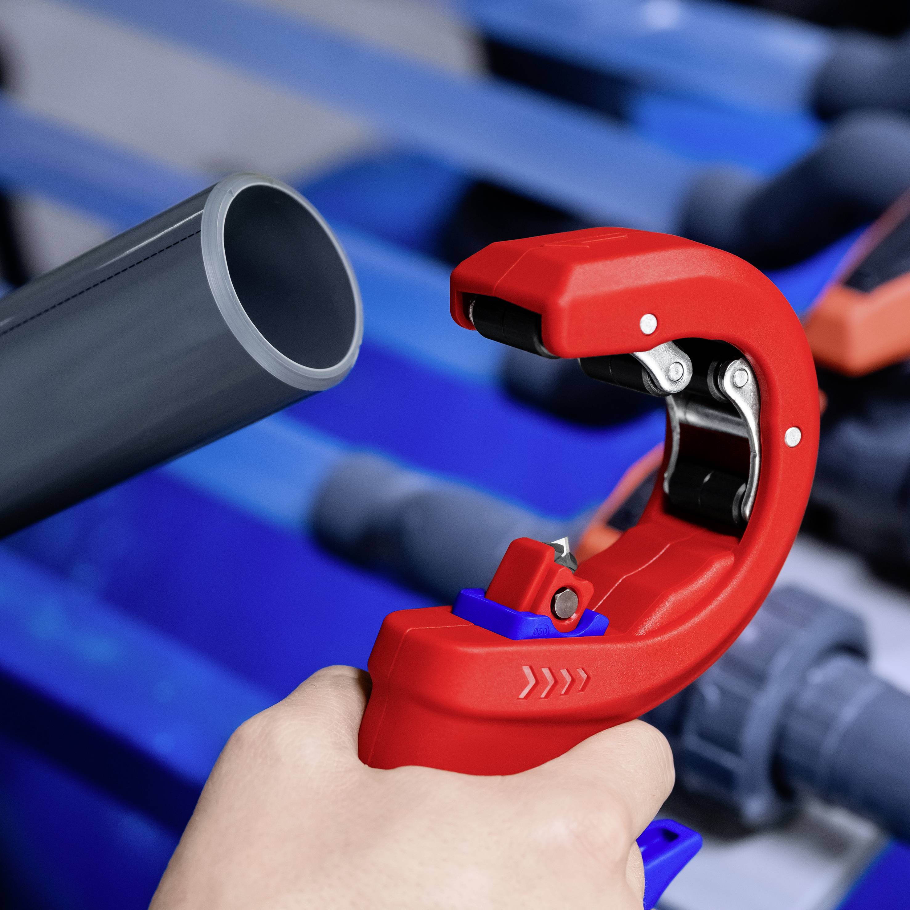 Rote Rohrschere schneidet graues Kunststoffrohr. Hintergrund unscharf, blaue und graue Töne dominieren. Arbeiterhand hält Rohr.