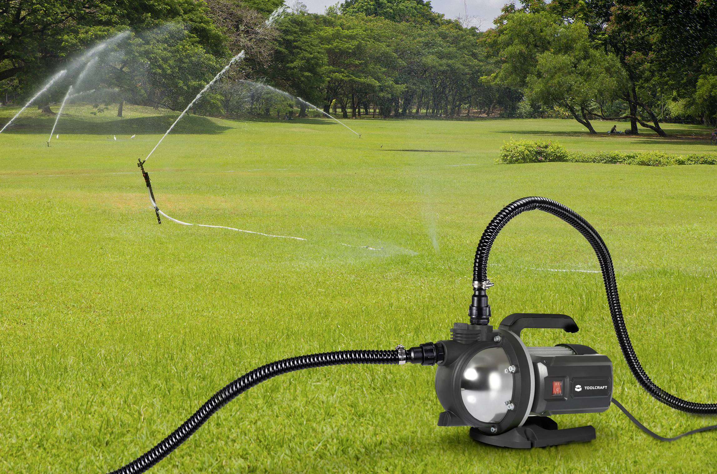 Gartenbewässerungssystem mit Sprinkler auf einer großen grünen Wiese, im Vordergrund eine schwarze Wasserpumpe, im Hintergrund Bäume.