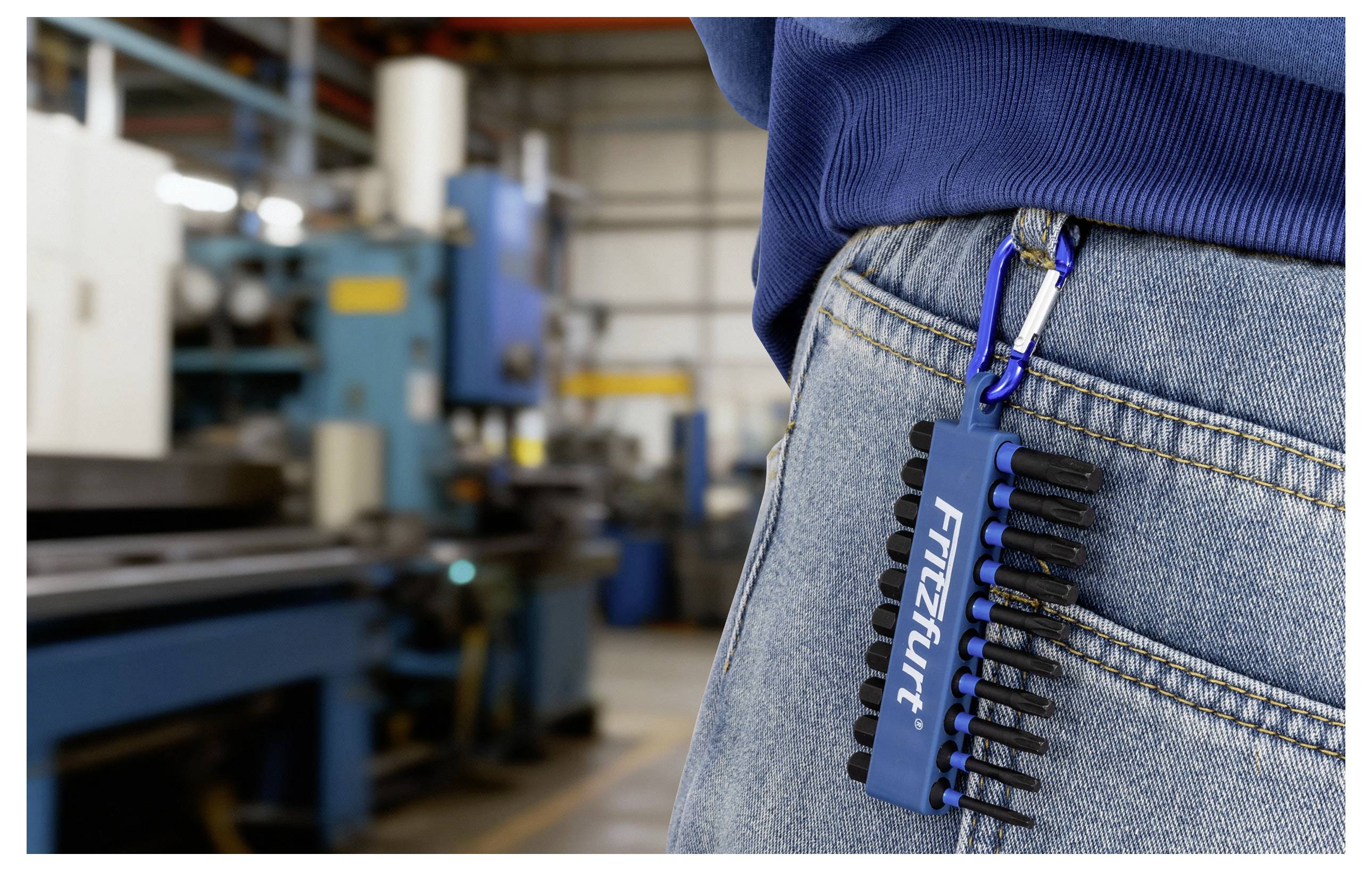 Arbeiter in Blue-Jeans mit einem seitlich angebrachten Werkzeughalterclip; Maschinen im Hintergrund einer Industriewerkstatt.