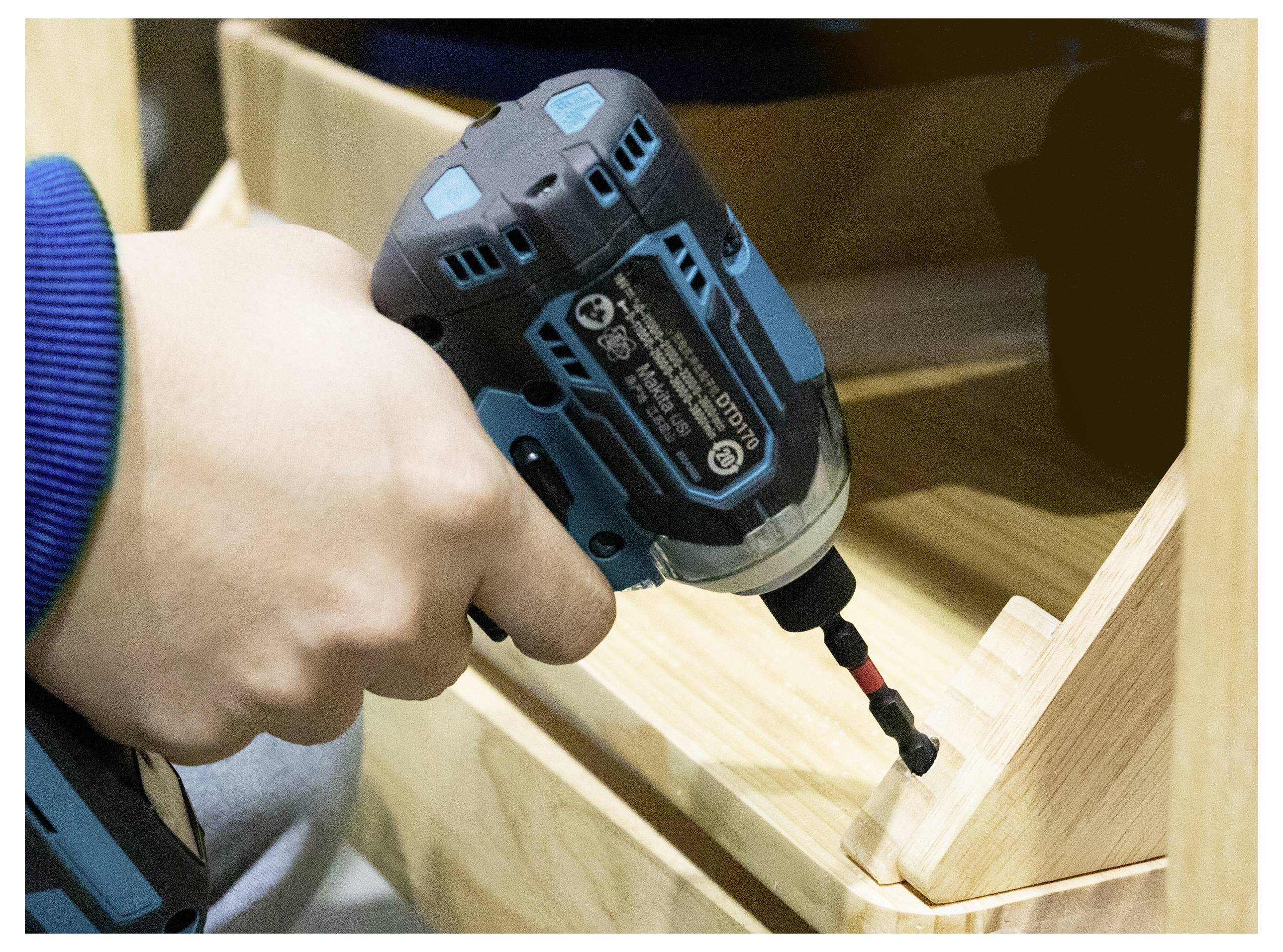 Eine Person verwendet einen blauen und schwarzen kabellosen Bohrer, um ein Holzmöbelstück zusammenzubauen und eine Schraube in eine Eckenverbindung einzudrehen.