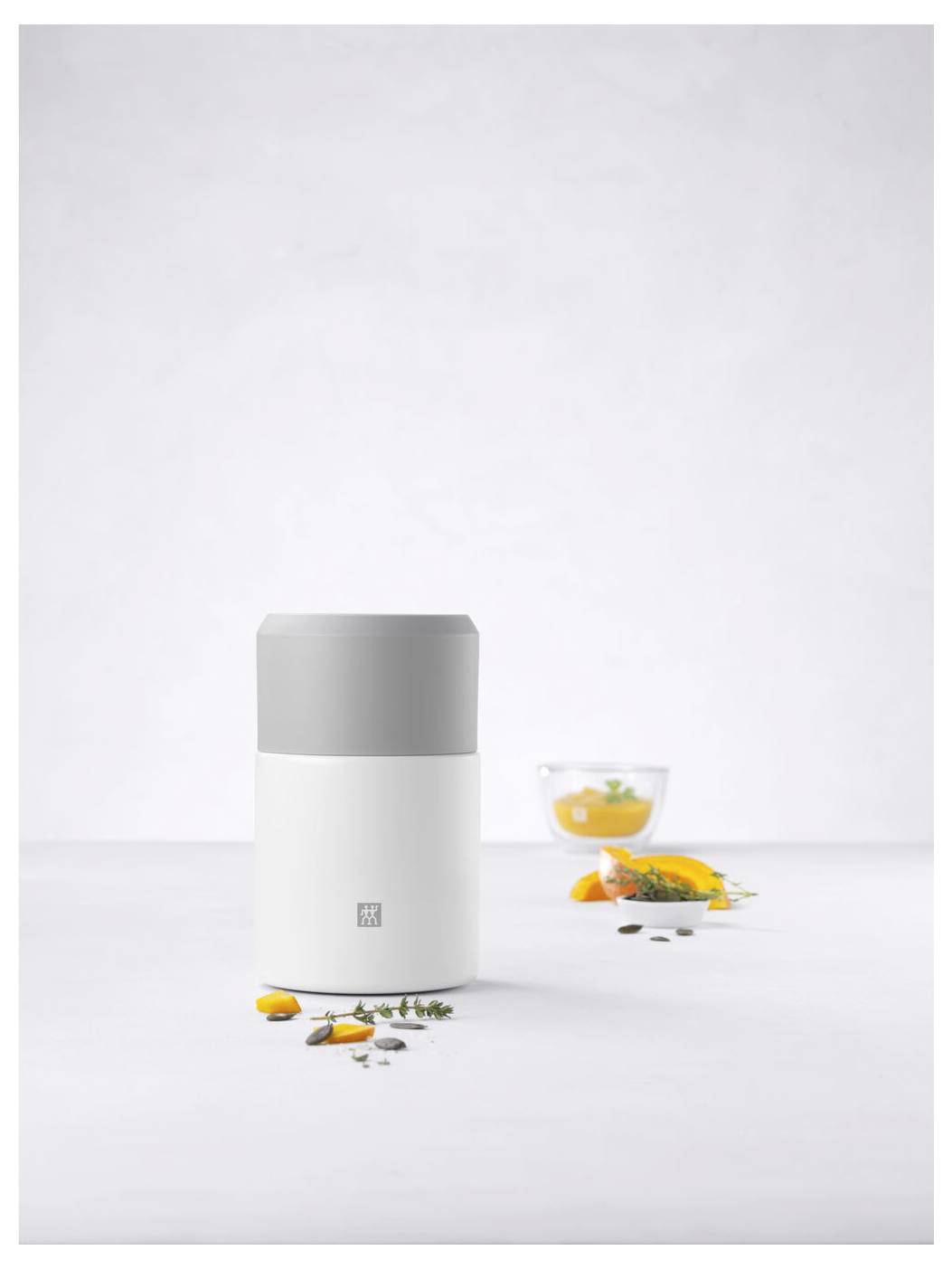 Küchenutensil mit grauem Deckel auf weißem Hintergrund, umgeben von Kräutern und Gewürzen. Im Hintergrund ein Glas mit Zitrone.