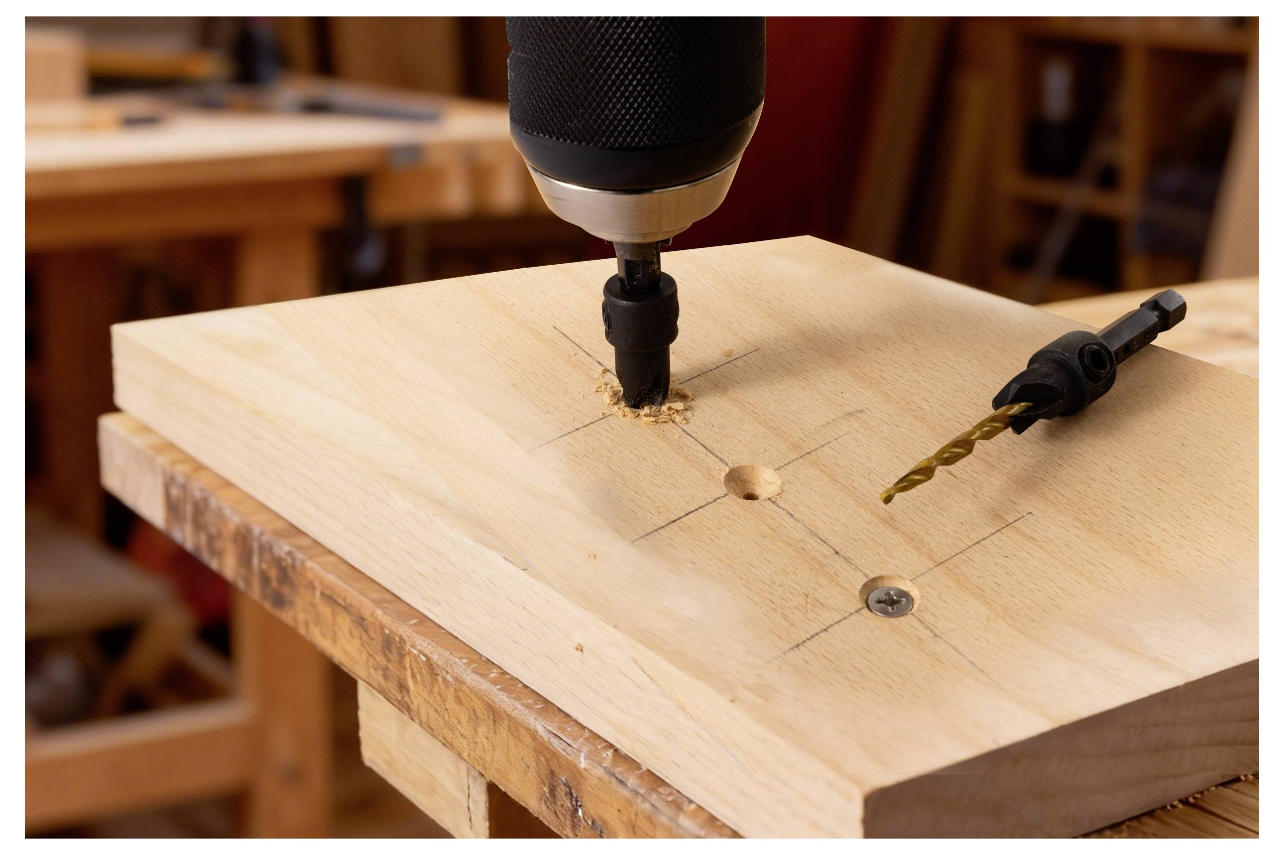 Ein Stück Holz, das auf einer Werkbank gebohrt wird. Ein Bohrer erzeugt Sägespäne um das Loch herum. Ein zusätzlicher Bohrer liegt in der Nähe.