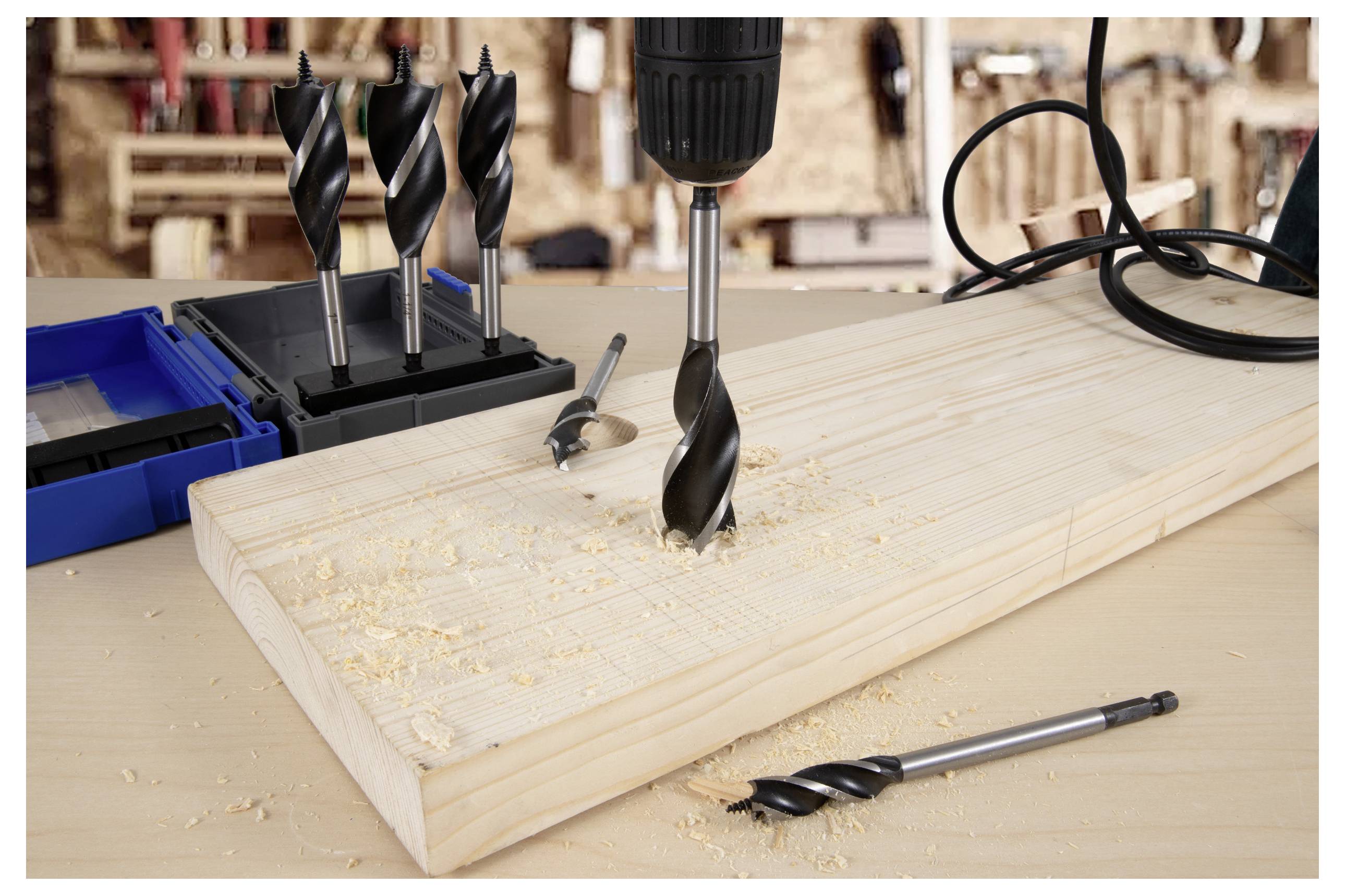 Eine Bohrmaschine mit einem großen Bohrer bohrt ein Loch in ein Holzbrett, umgeben von Sägespänen und anderen Bohrern auf einer Werkbank.