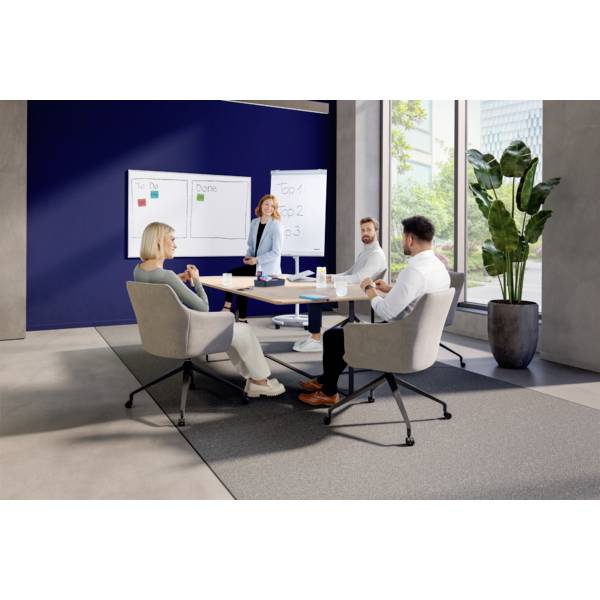 Eine Frau steht vor einem Whiteboard und präsentiert. Drei Personen sitzen und hören zu. Im Raum sind Pflanzen und große Fenster.