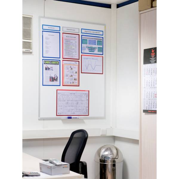 Ein Whiteboard hängt an der Wand, mit bunten Plakaten und Diagrammen. Davor steht ein schwarzer Bürostuhl, daneben ein Mülleimer.