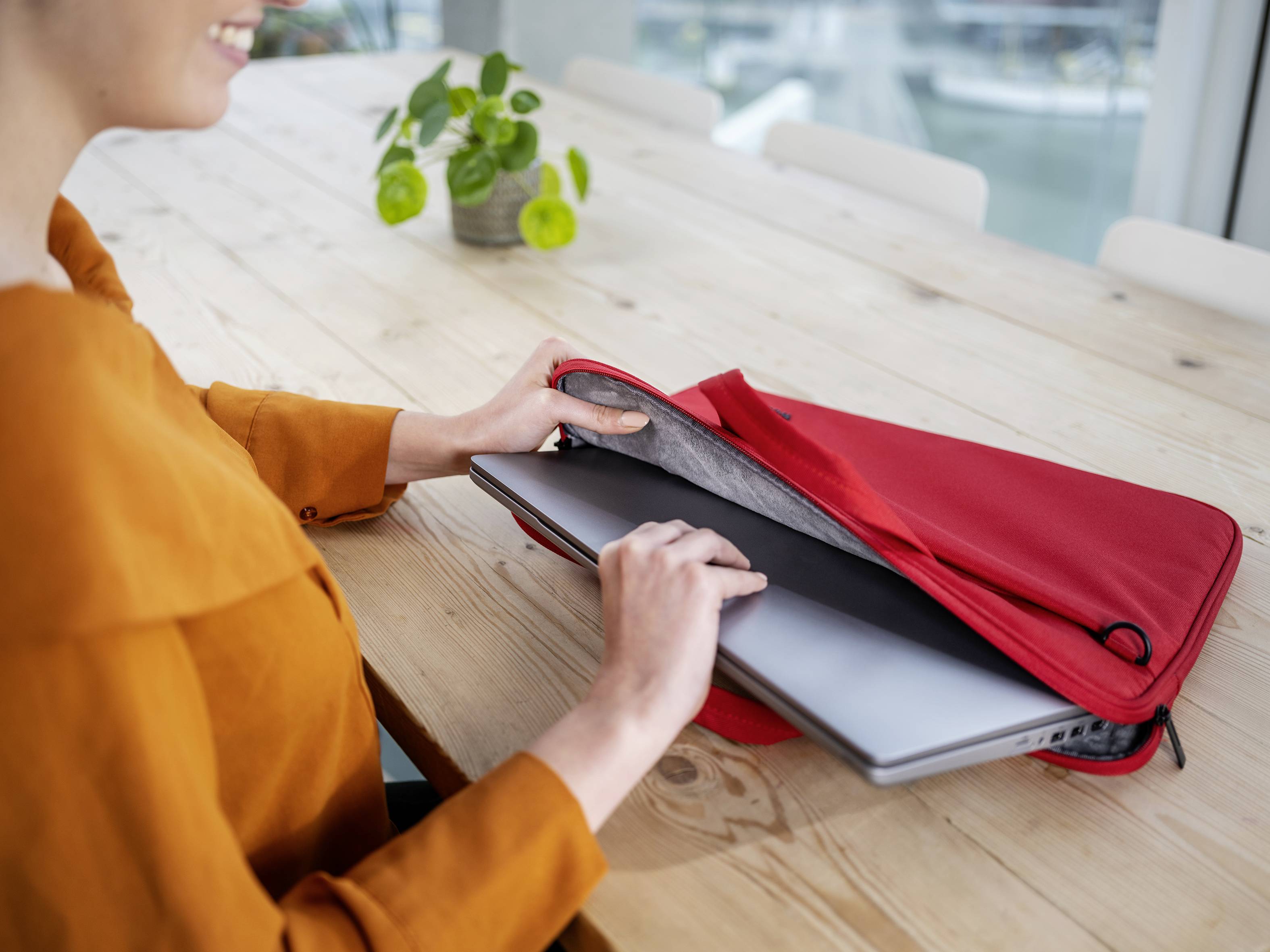 Eine Person steckt einen Laptop in eine rote Tasche auf einem Holztisch. Im Hintergrund steht eine kleine Pflanze.