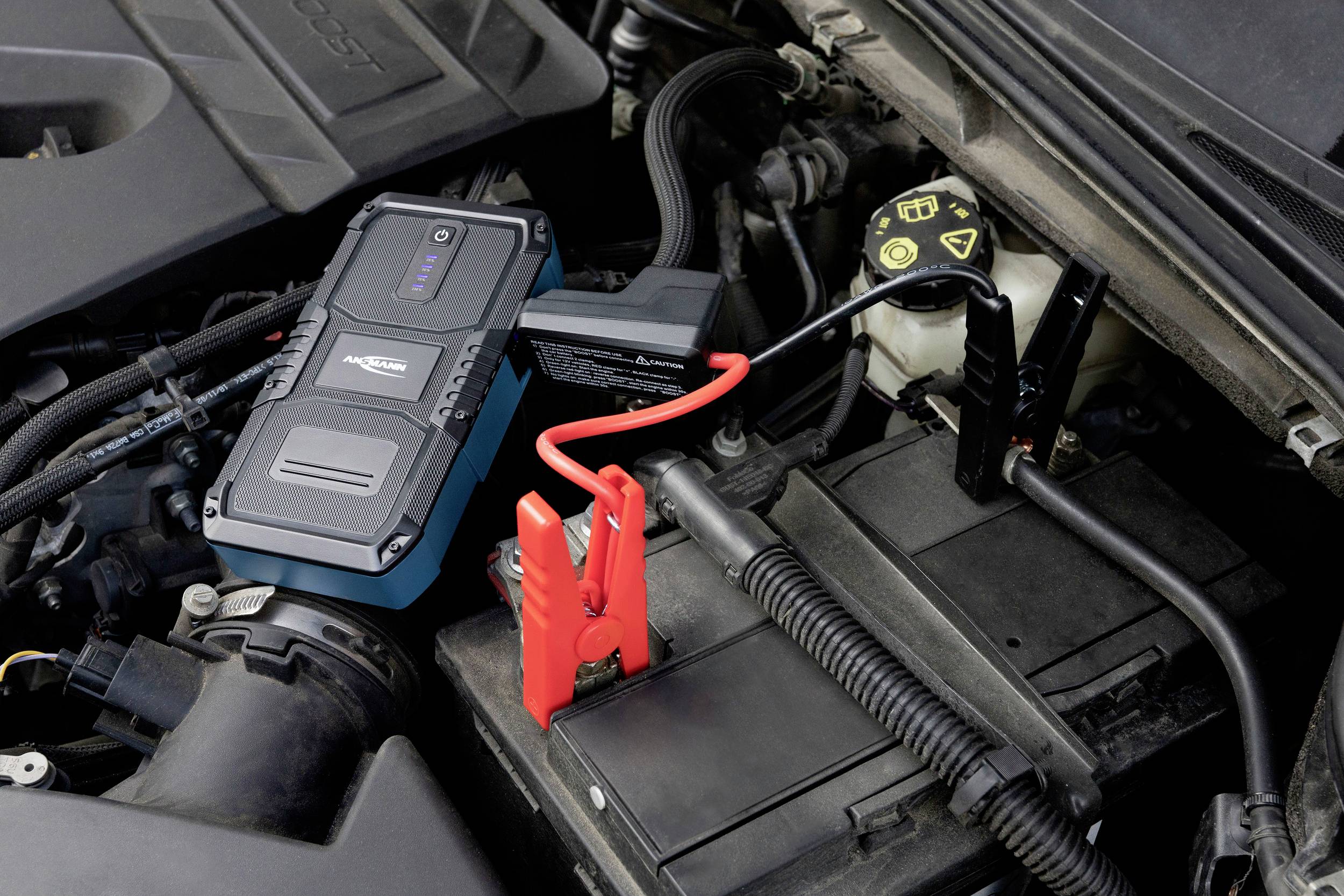 Ein Starthilfegerät ist an eine Autobatterie angeschlossen. Ein rotes Überbrückungskabel verbindet das Gerät mit dem Pluspol der Batterie.