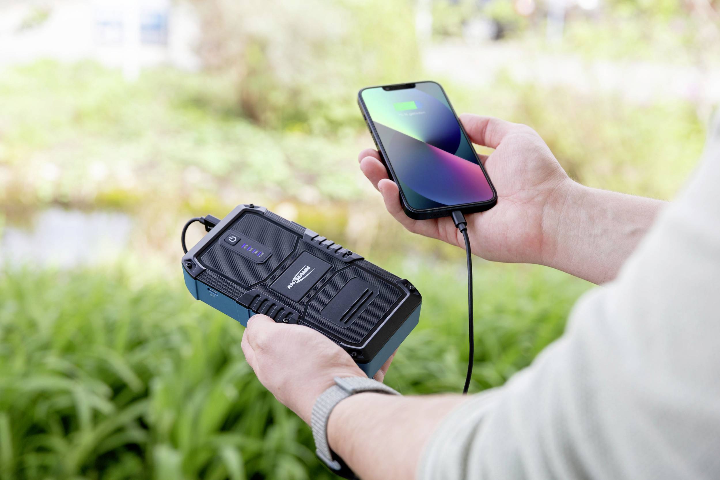 Person lädt draußen ein Smartphone mit einem tragbaren Akku auf; der Akku zeigt volle Ladung an.