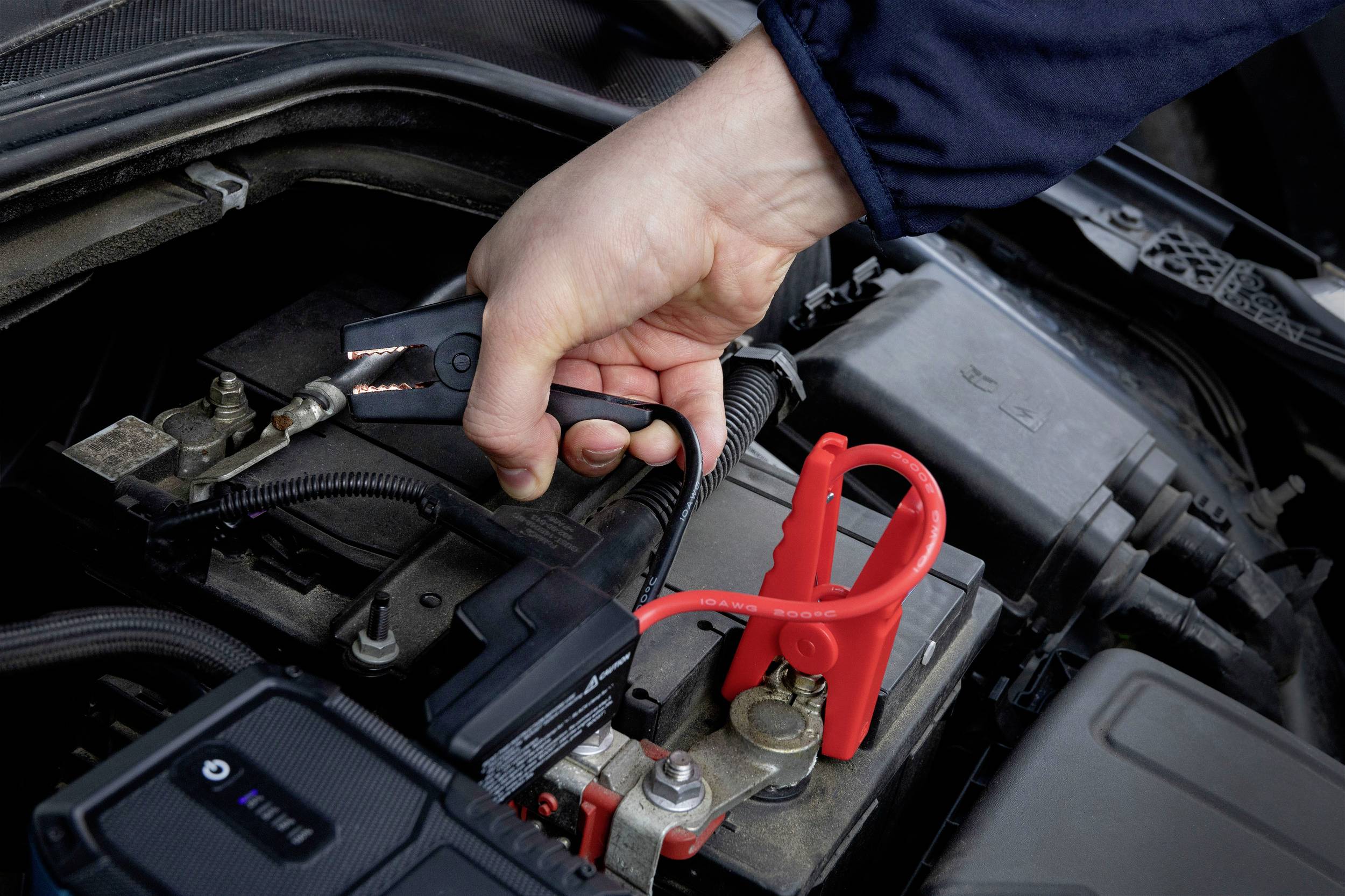 Eine Hand schließt ein schwarzes Starthilfekabel an die Batterie eines Autos an, um die Fahrzeugbatterie aufzuladen.
