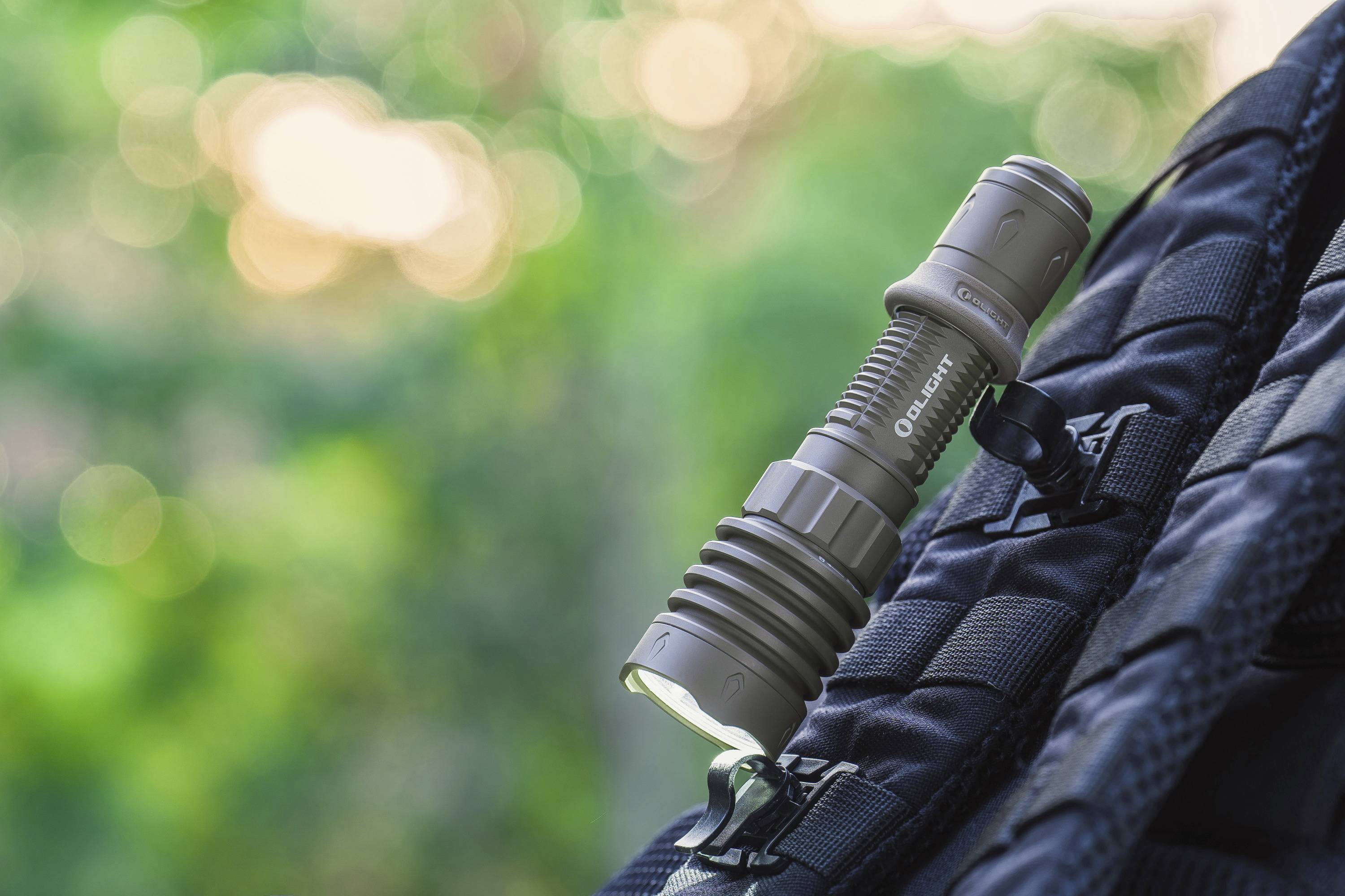 Eine graue Taschenlampe ist an einem schwarzen Rucksack befestigt. Der Hintergrund zeigt verschwommene, grüne Natur.