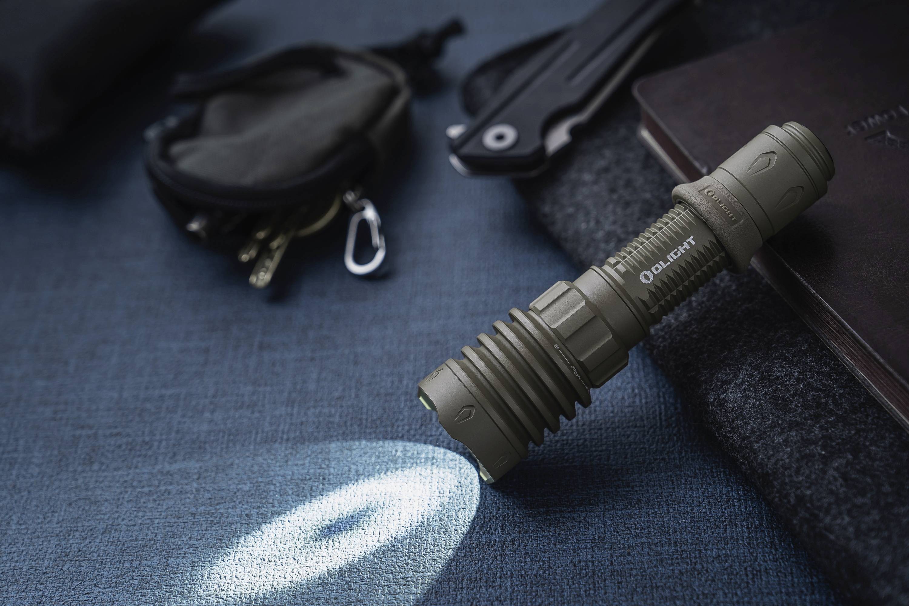 Eine Taschenlampe leuchtet auf einem blauen Stoffuntergrund. Daneben liegen ein Messer, ein Schlüsselbund und eine Tasche.