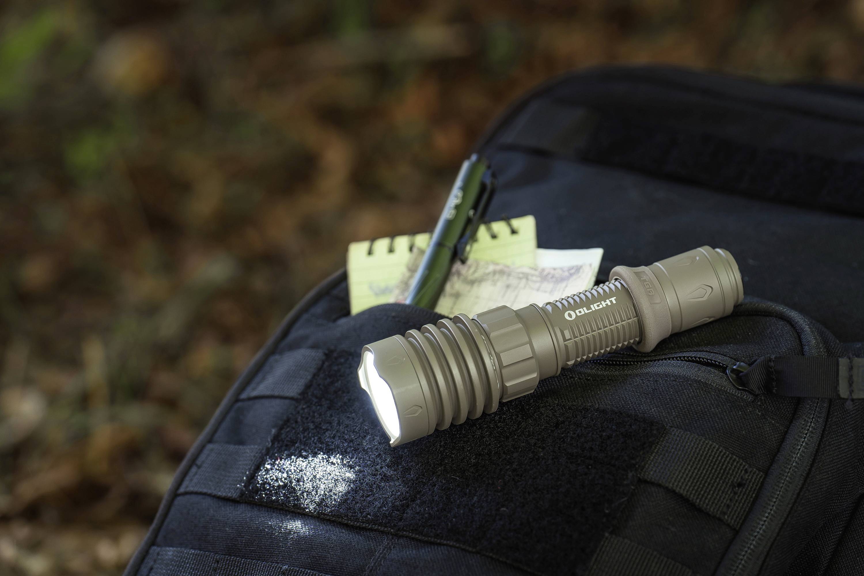 Eine Taschenlampe liegt eingeschaltet auf einem schwarzen Rucksack neben einem Notizblock und einem Stift im Freien.