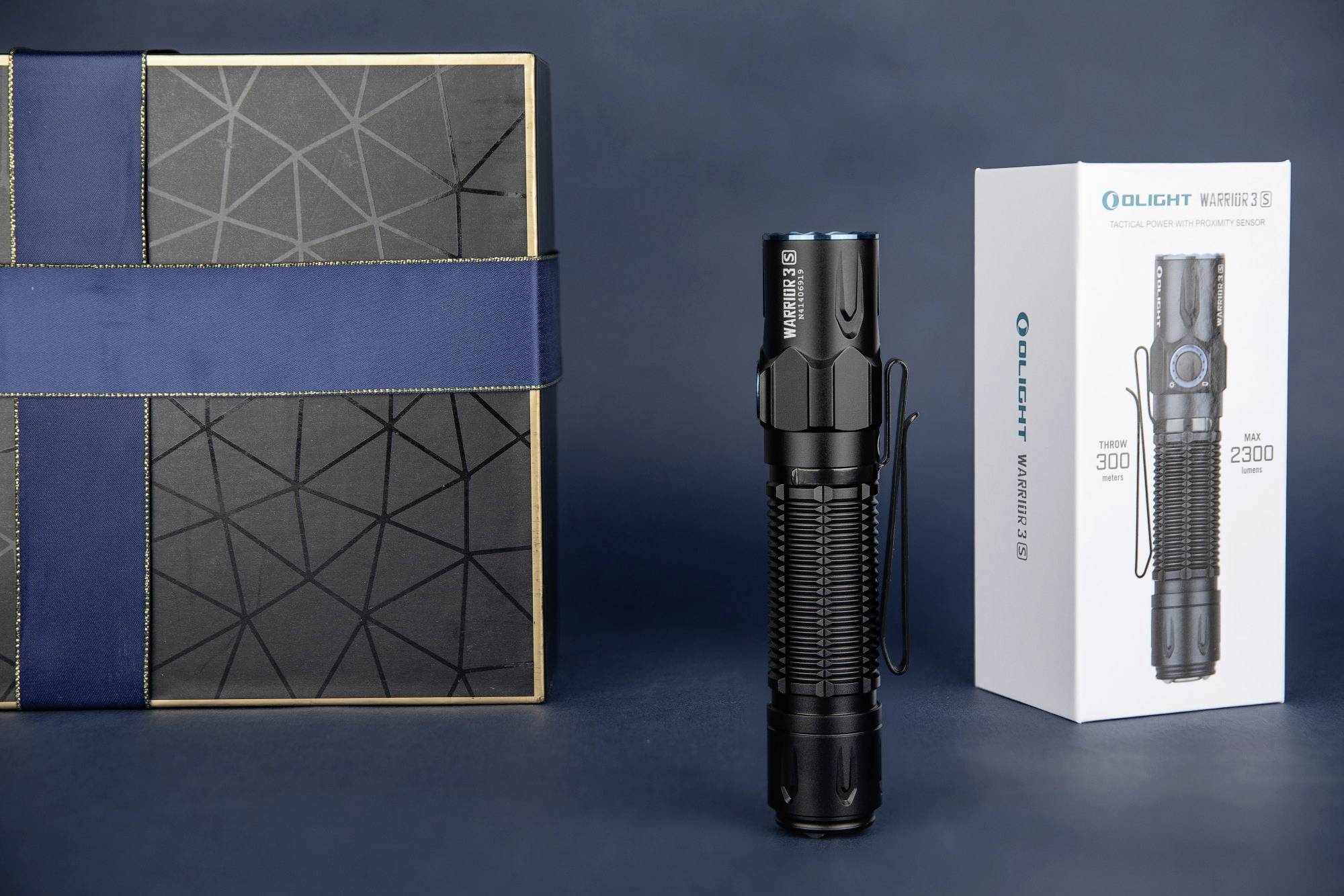 Eine schwarze Taschenlampe steht aufrecht zwischen einer dekorativen Box und einer Verpackung mit der Aufschrift 'Olight Warrior 3S'.