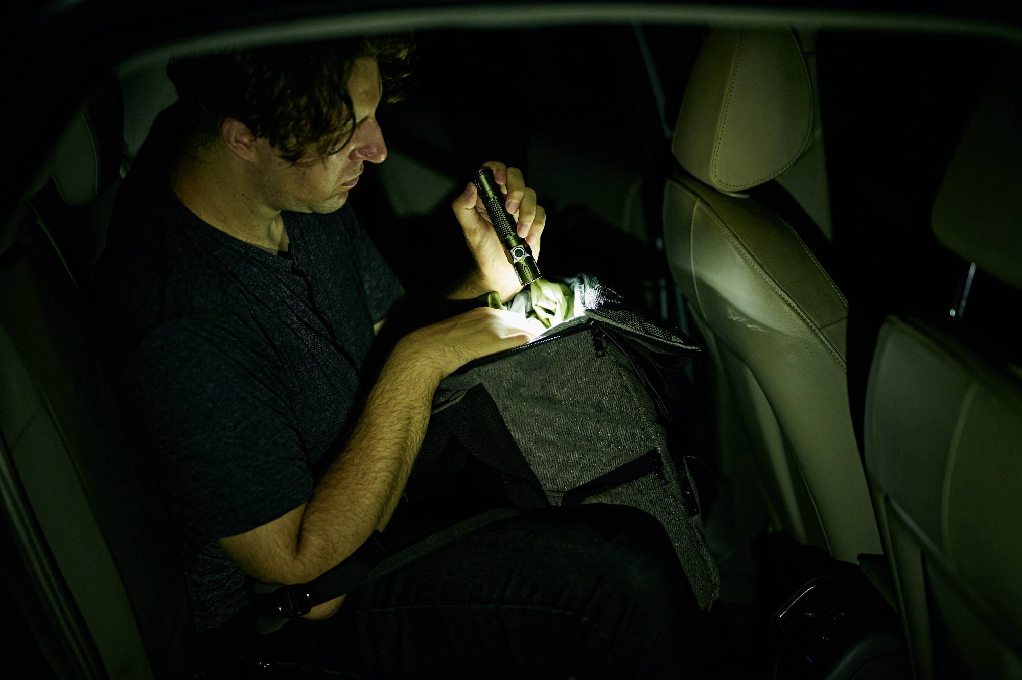 Ein Mann sitzt im Auto und leuchtet mit einer Taschenlampe in eine Tasche. Es ist dunkel, und er scheint etwas zu suchen.