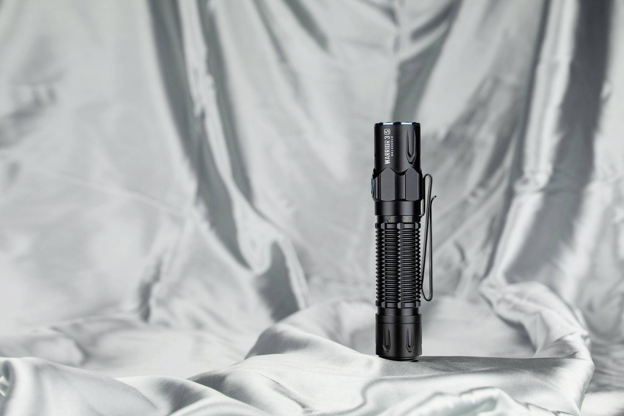 Schwarze Taschenlampe steht auf silbernem, glänzendem Stoffhintergrund. Sie hat einen Clip an der Seite und wirkt robust.