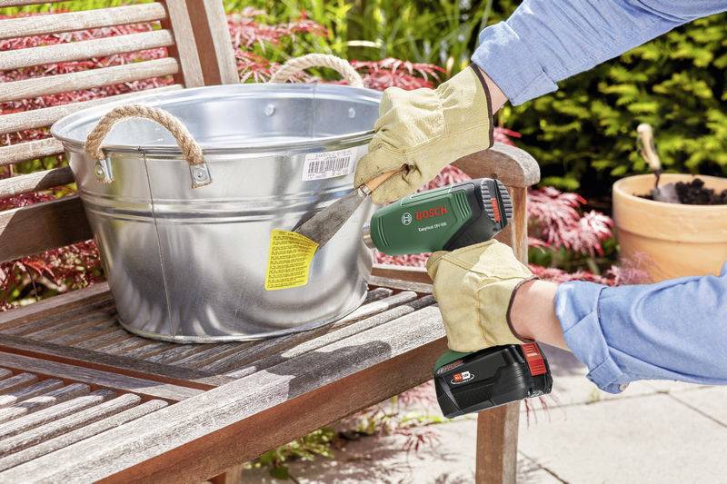 Person entfernt mit einem Bosch-Akku-Schaber ein Etikett von einem silbernen Eimer, der auf einer Holzbank im Garten steht.