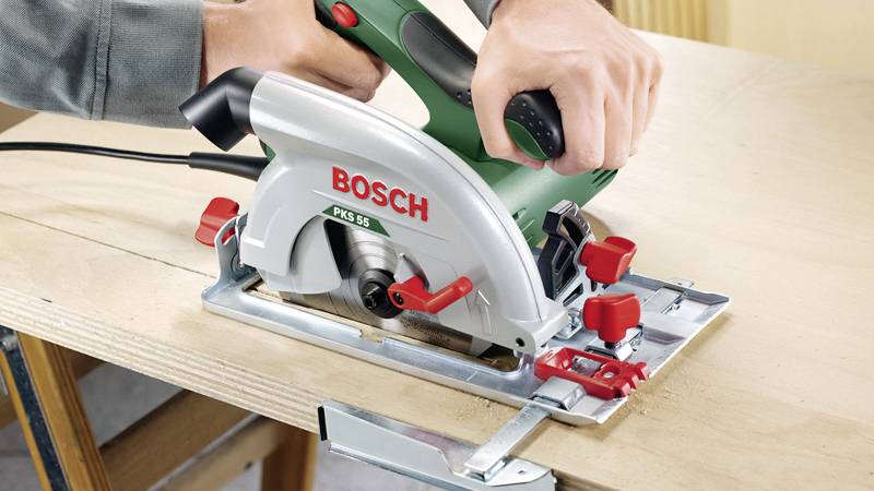 Eine Hand hält eine grüne elektrische Kreissäge der Marke 'Bosch' und schneidet präzise durch ein Holzbrett.