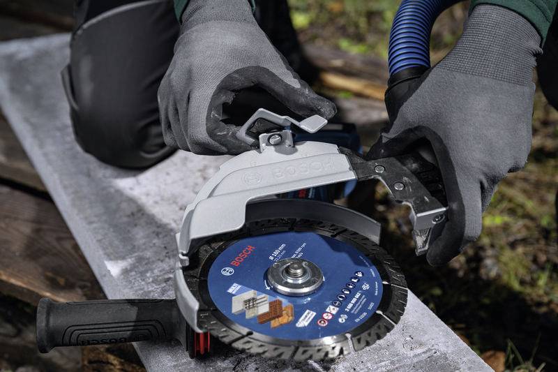 Eine Person mit grauen Handschuhen befestigt eine Schutzabdeckung an einer blauen Bosch-Trennscheibe, die auf einem Metallträger liegt.